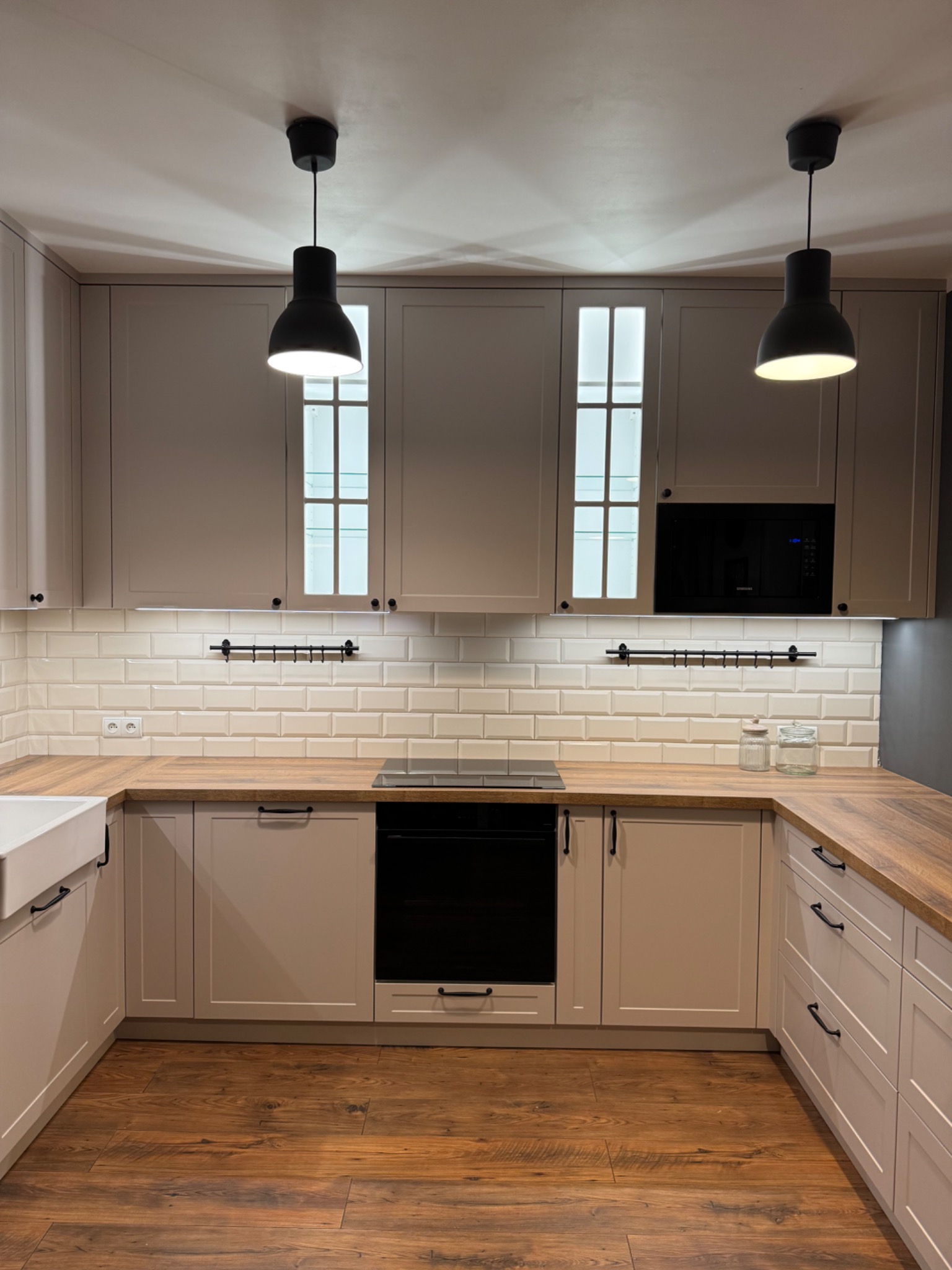 Jasna kuchnia z drewnianym blatem, białymi płytkami na ścianie i czarnymi lampami wiszącymi, widok na zlew i płytę grzewczą.