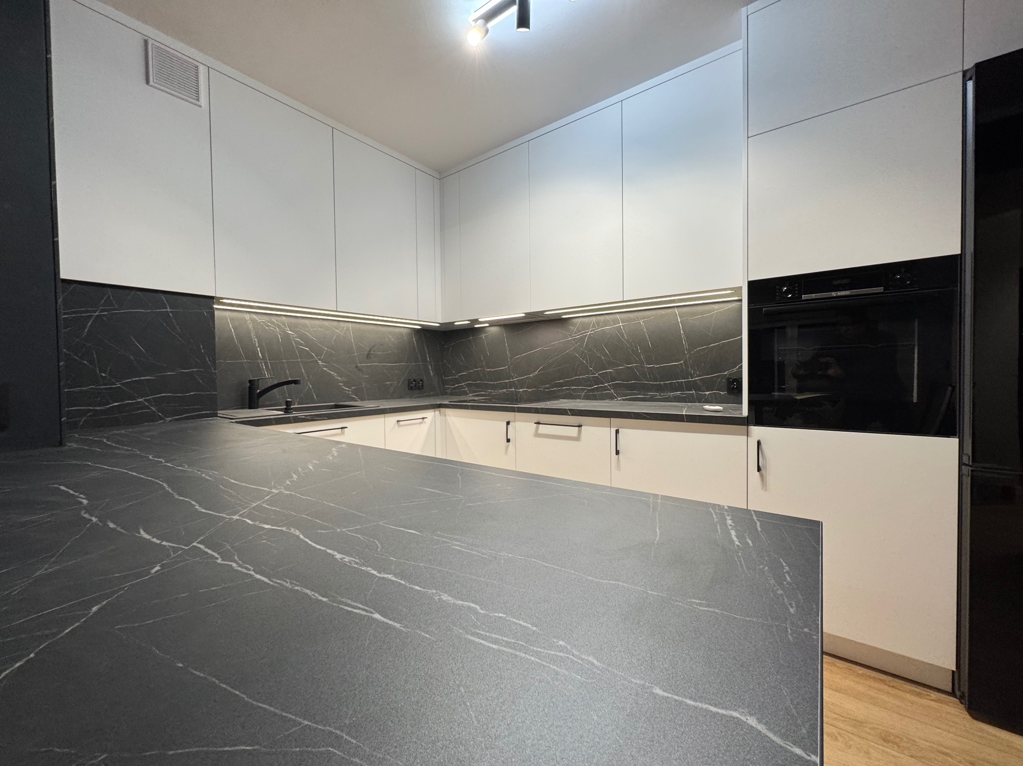 Nowoczesna kuchnia z białymi szafkami i blatem imitującym czarny marmur z białymi żyłami, oświetlenie LED podszafkowe, czarny piekarnik w zabudowie.