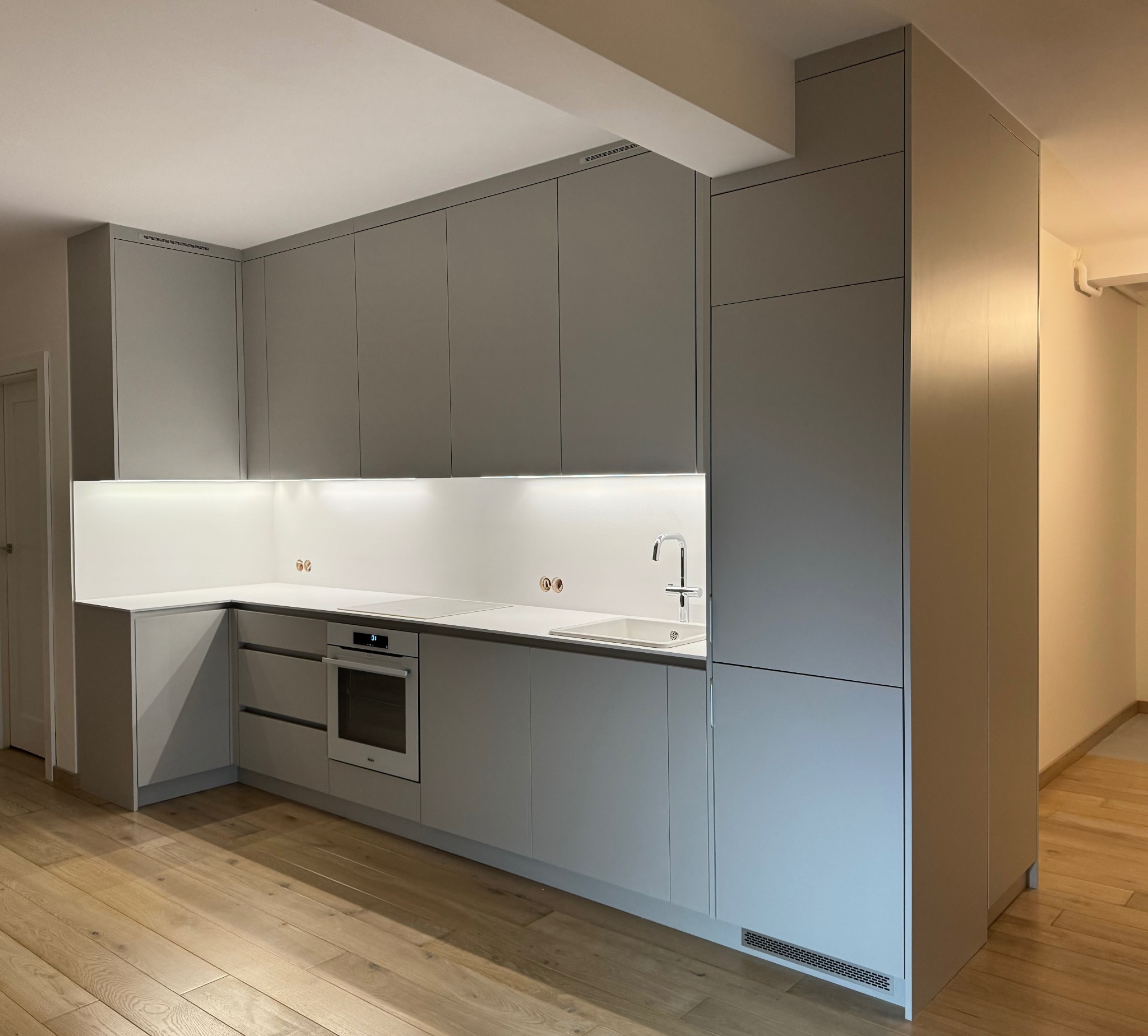 Nowoczesna kuchnia w odcieniach szarości z drewnianą podłogą, oświetleniem LED pod szafkami i minimalistyczną armaturą.