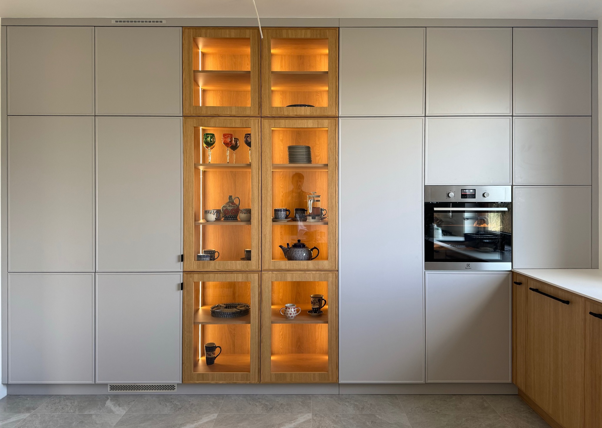 Nowoczesna kuchnia na wymiar w Radwanicach z szarymi frontami i przeszklonymi witrynami podświetlonymi ciepłym światłem, eksponującymi kolekcję naczyń i dekoracyjnych przedmiotów.