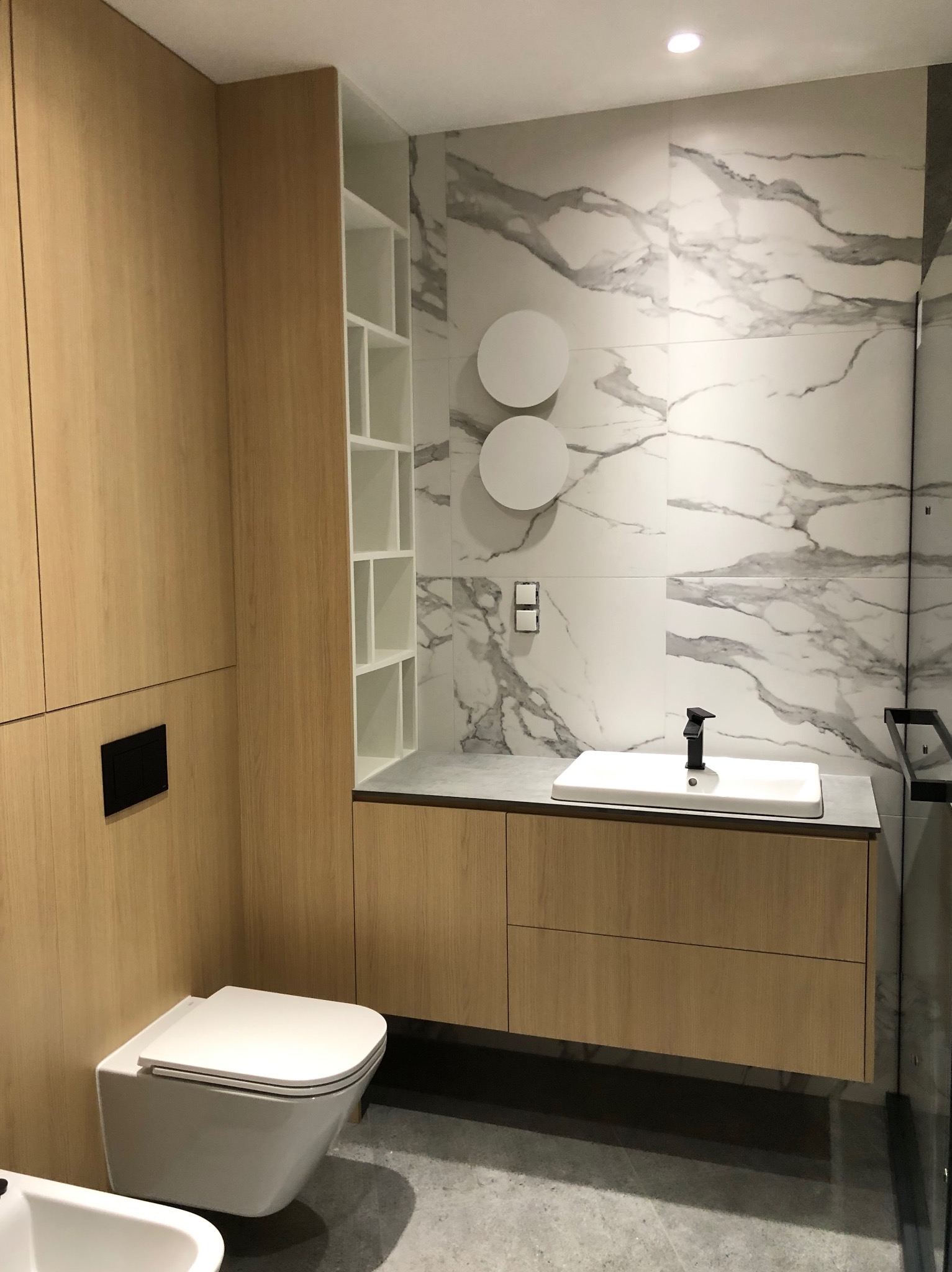 Nowoczesna łazienka z zabudową z jasnego drewna, marmurowymi płytkami na ścianie i wiszącą toaletą.