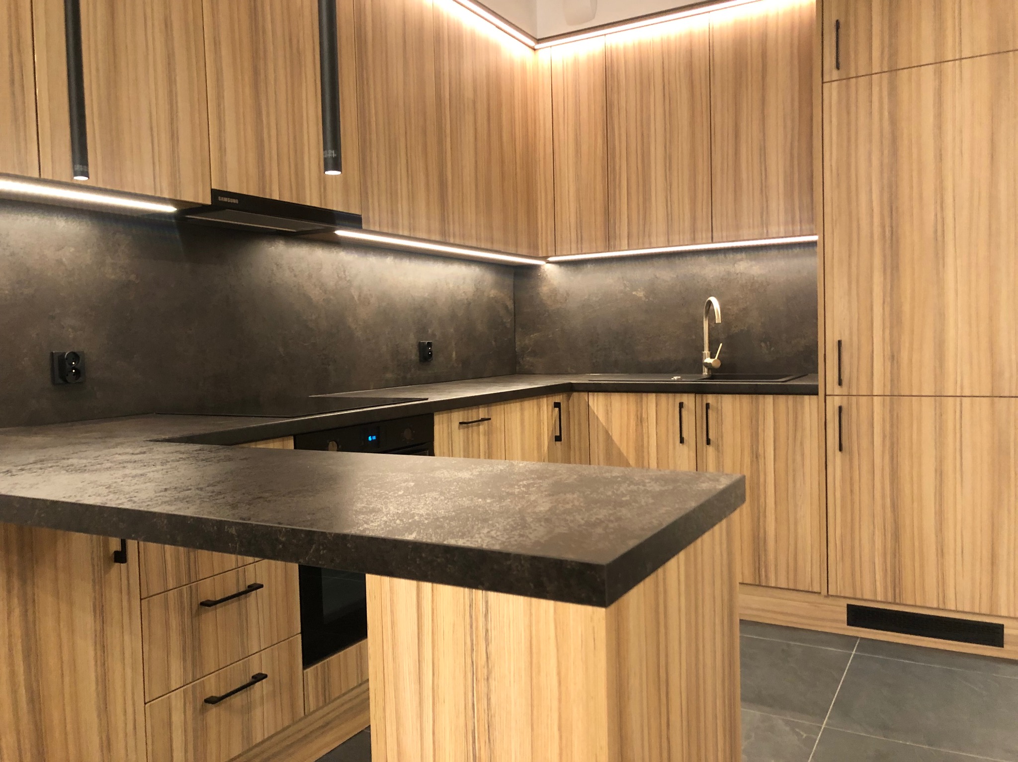 Nowoczesna kuchnia z drewnianymi frontami szafek o wyraźnym usłojeniu, ciemnym blatem i podświetleniem LED podszafkowym oraz punktowymi lampami sufitowymi.