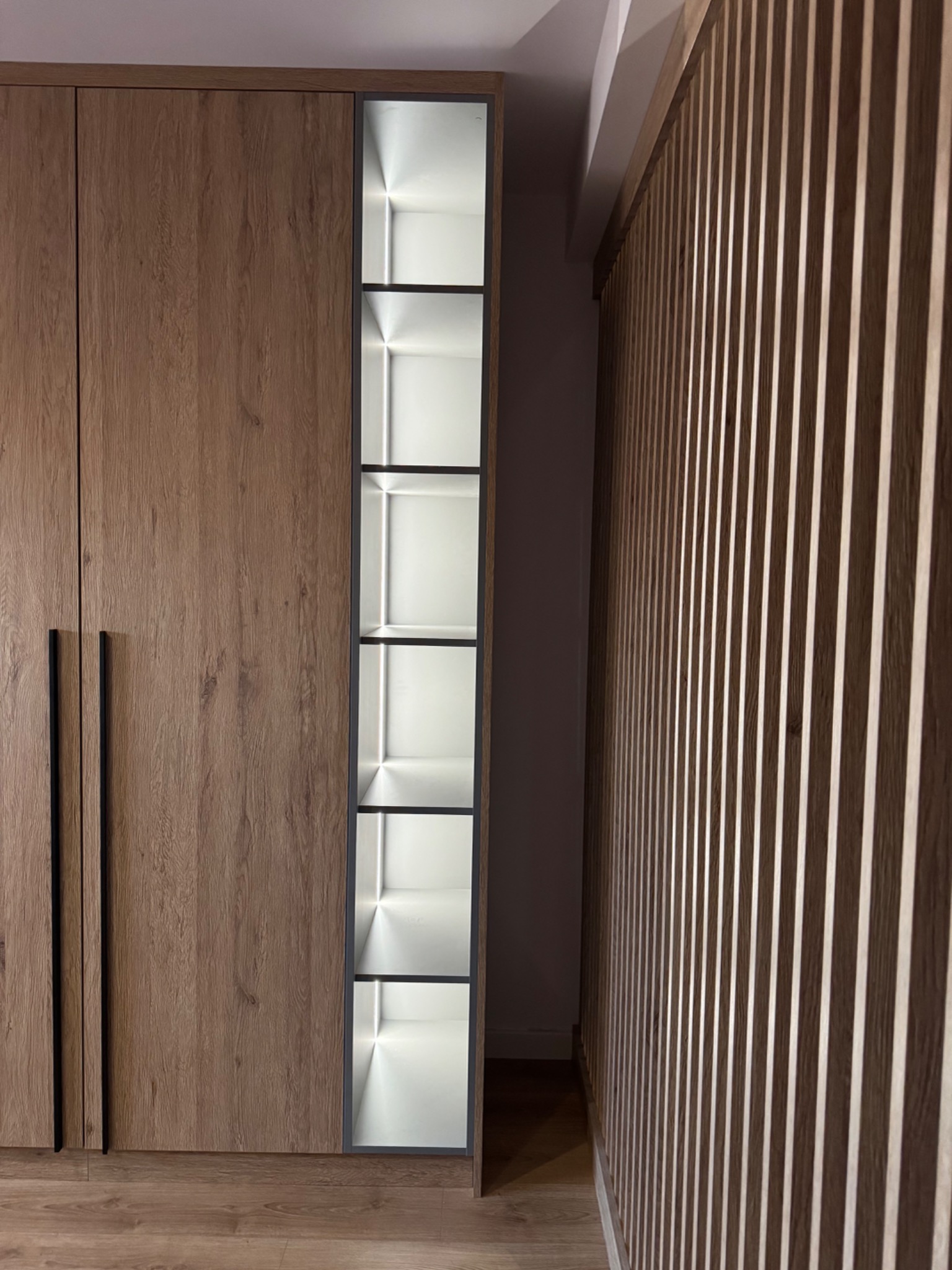 Drewniana szafa z czarnymi uchwytami i wbudowanymi, podświetlonymi półkami, obok dekoracyjna ściana z drewnianych listewek.