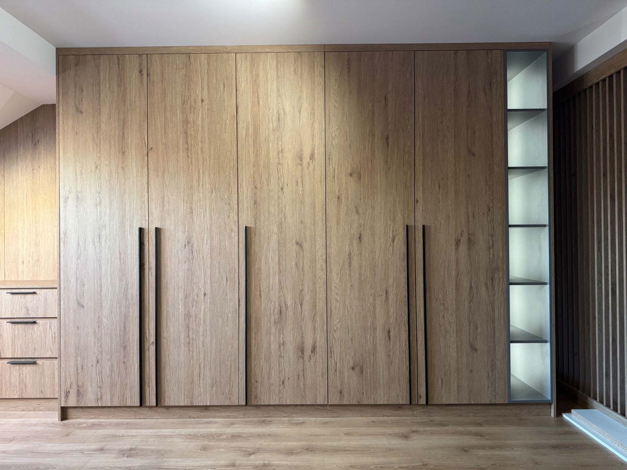 Duża szafa wnękowa z drewnianymi frontami i czarnymi uchwytami, z otwartym regałem z podświetlanymi półkami po prawej stronie, dopasowana do drewnianej podłogi.