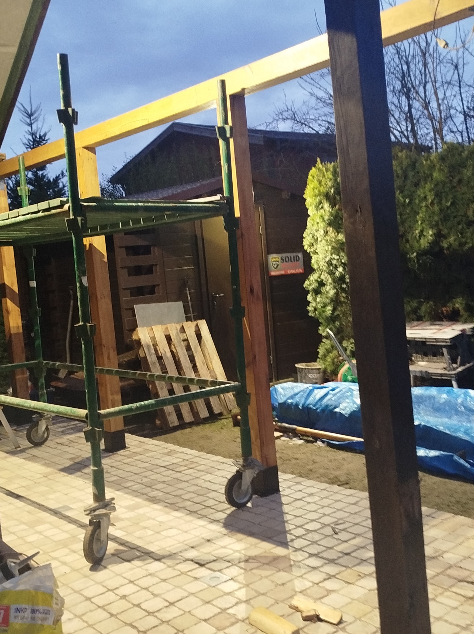 Konstrukcja z drewnianych belek na zewnątrz, widoczne rusztowanie, palety, plandeka i budynek w tle.