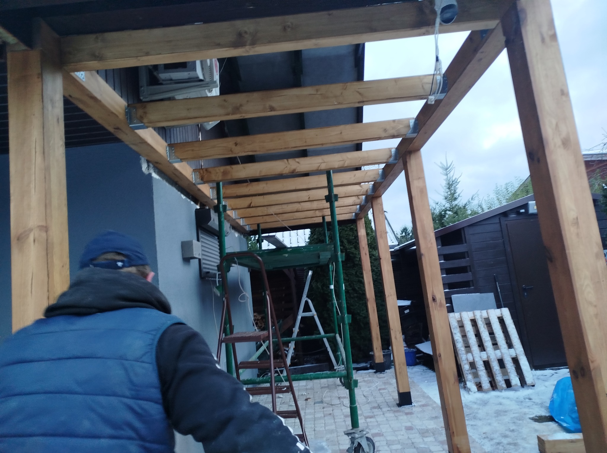 Konstrukcja drewnianej pergoli w trakcie montażu, widoczny pracownik na rusztowaniu, drewniane belki, metalowe łączniki oraz paleta z resztkami śniegu w tle.