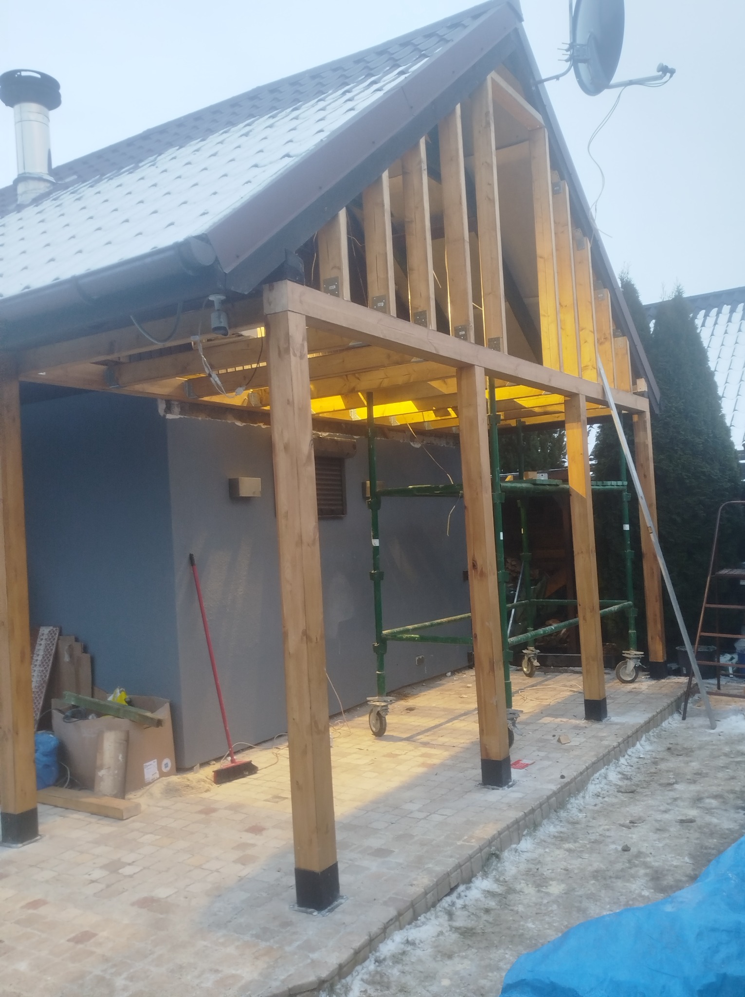 Konstrukcja drewniana z elementami szkieletu dachu w trakcie budowy przy budynku z szarą elewacją, widoczne rusztowania i materiały budowlane w zimowej scenerii.