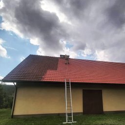 MK ULTRA - Pracownik w kombinezonie na drabinie czyści prawą stronę dachu z czerwonej dachówki, widoczna różnica w kolorze między czystą a zabrudzoną częścią, na tle pochmurnego nieba.