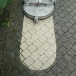 Czyszczenie kostki brukowej za pomocą okrągłej myjki ciśnieniowej; widoczny kontrast między czystą, jasną powierzchnią a brudną, ciemną kostką.