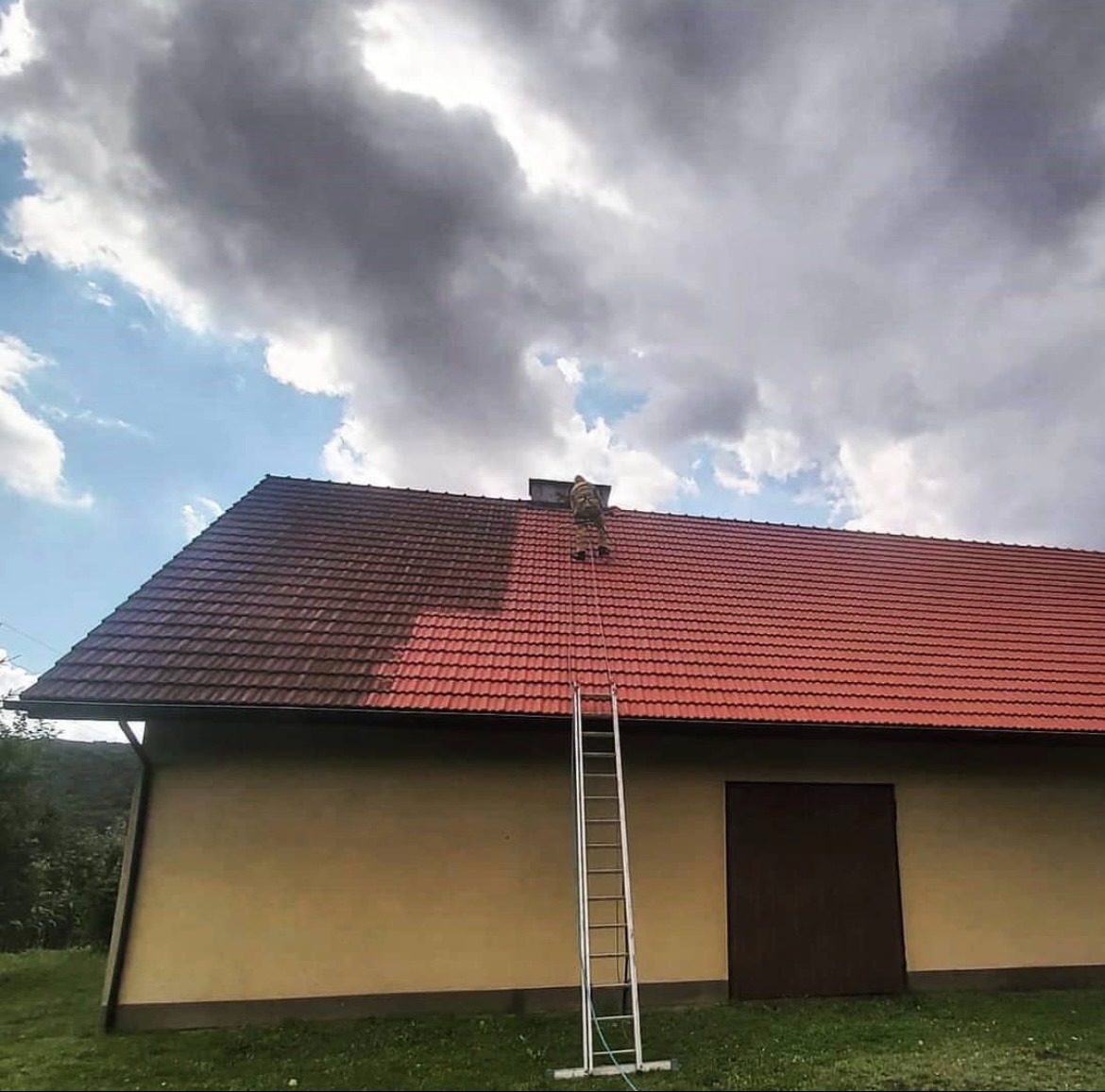 Pracownik w kombinezonie na drabinie czyści prawą stronę dachu z czerwonej dachówki, widoczna różnica w kolorze między czystą a zabrudzoną częścią, na tle pochmurnego nieba.