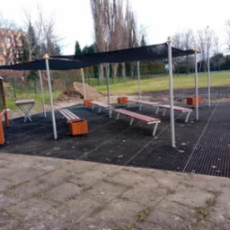 Zadaszone miejsce odpoczynku z ławkami i donicami na ażurowej platformie, obok widoczna pryzma ziemi i chodnik z kostki.