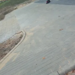 Ułożona szara kostka brukowa na podjeździe, widoczna krawędź z obrzeży, a obok wzmocnione siatką skarpy ziemne.