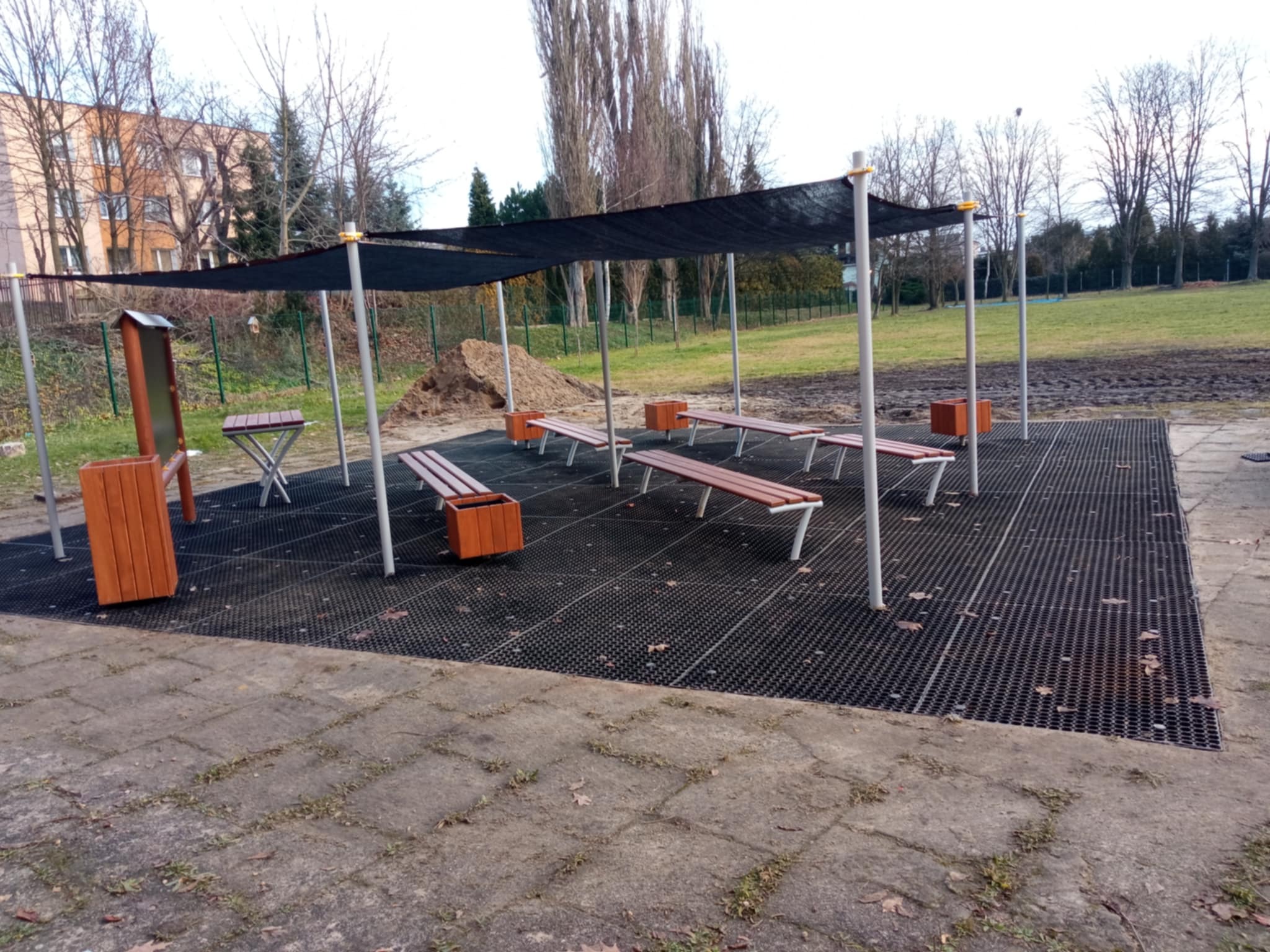 Zadaszone miejsce odpoczynku z ławkami i donicami na ażurowej platformie, obok widoczna pryzma ziemi i chodnik z kostki.