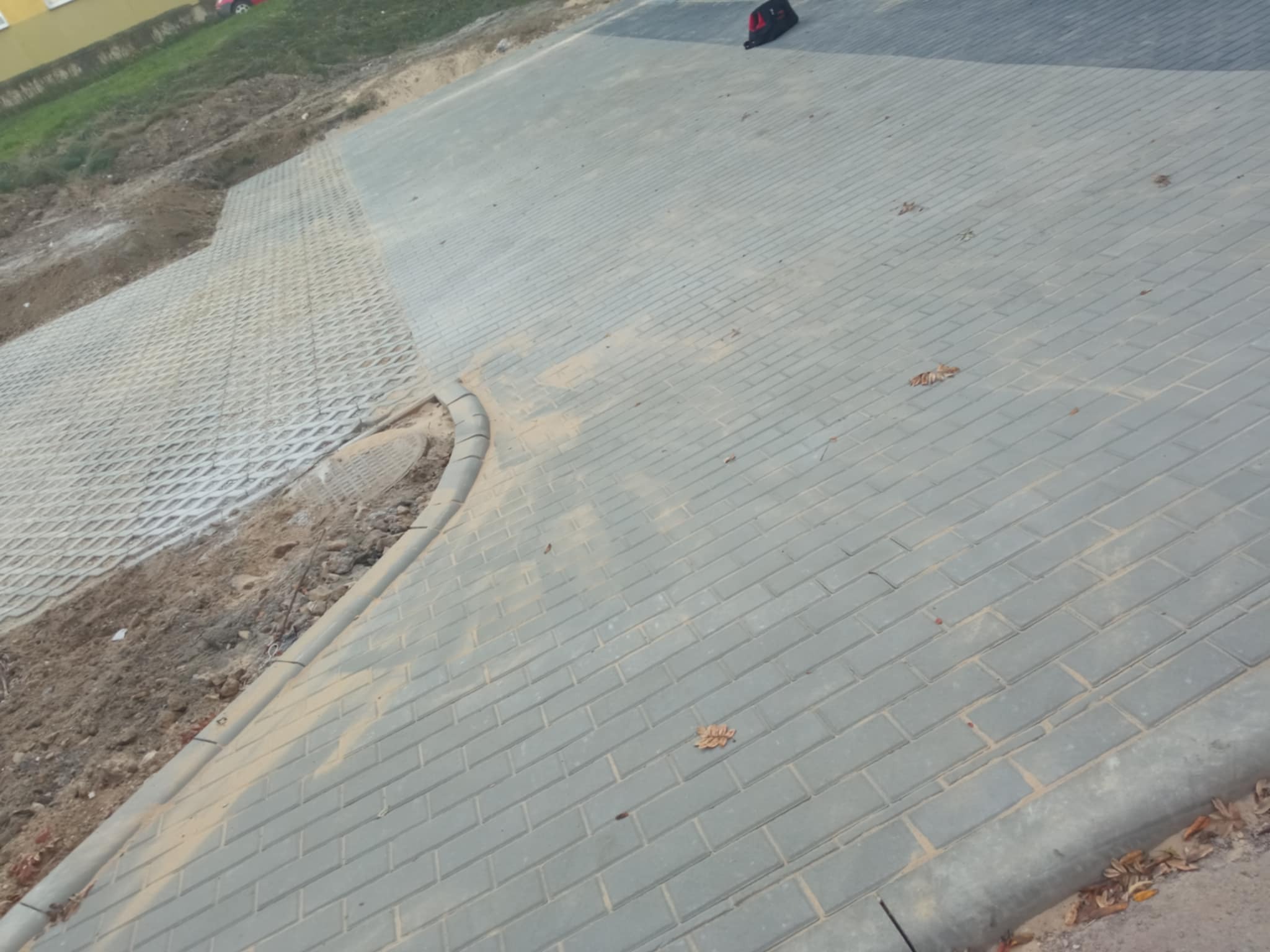 Ułożona szara kostka brukowa na podjeździe, widoczna krawędź z obrzeży, a obok wzmocnione siatką skarpy ziemne.