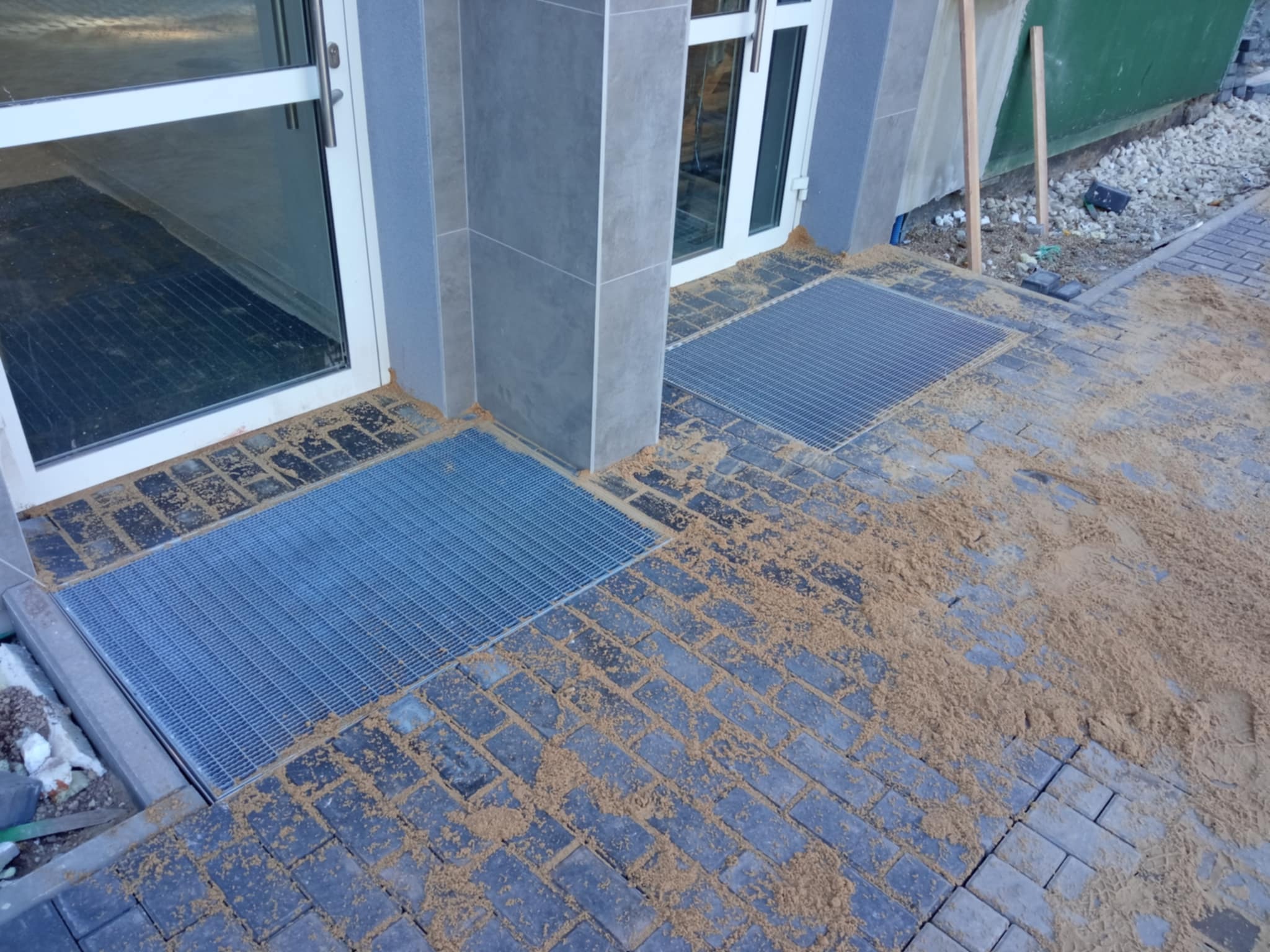 Ułożona kostka brukowa w odcieniach szarości z kratkami odpływowymi przy wejściu do budynku, posypana piaskiem.