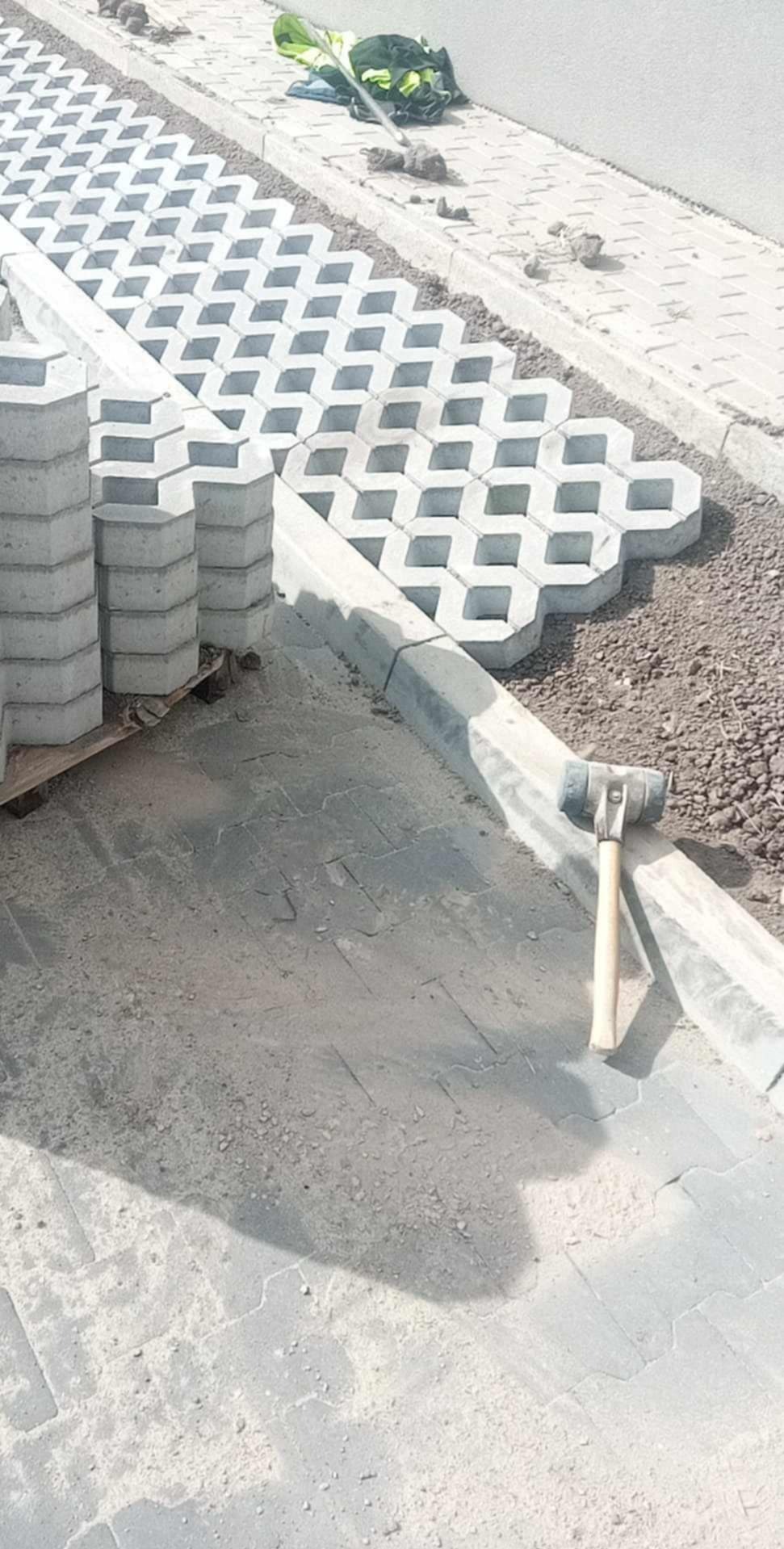 Ułożone ażurowe płyty betonowe na podsypce piaskowej, obok stos kostki brukowej i gumowy młotek brukarski.