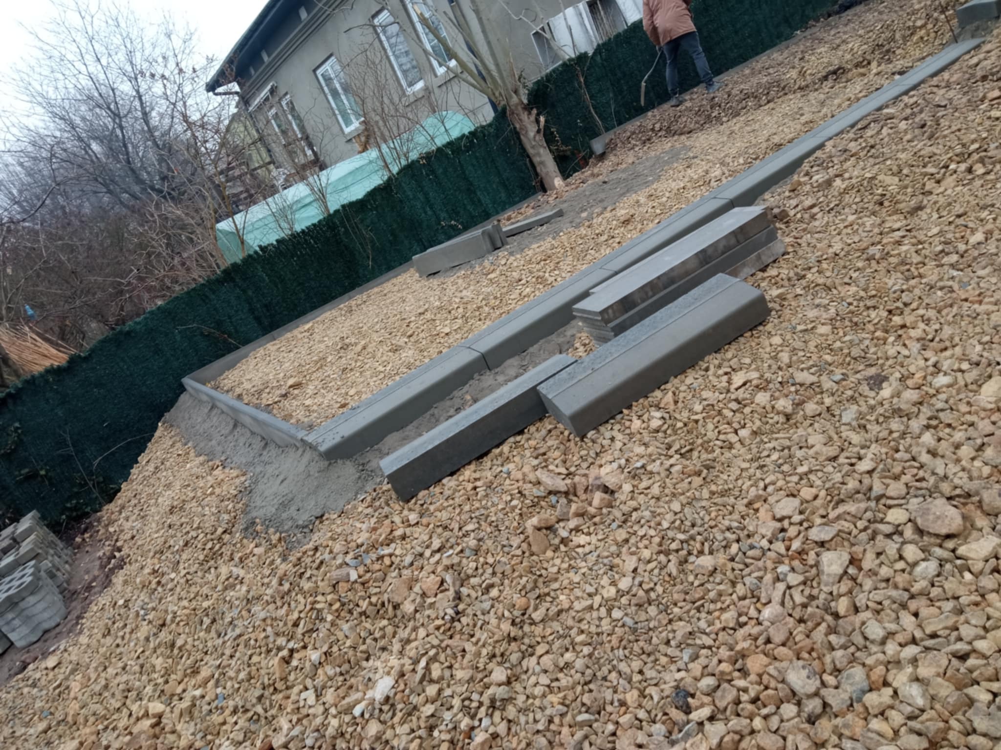 Przygotowanie terenu pod ścieżkę z obrzeżami betonowymi na żwirowym podłożu, widoczny fragment budynku i siatka ogrodzeniowa w tle.