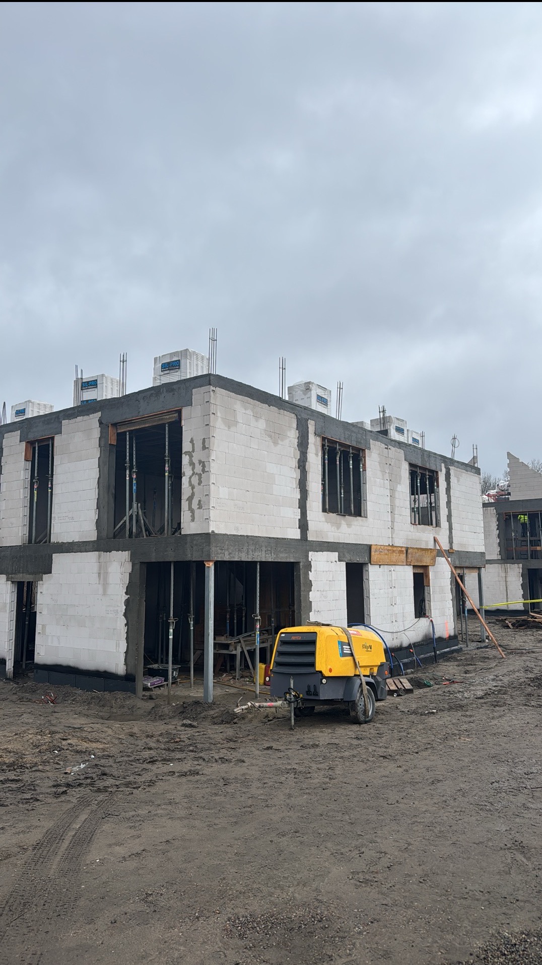 Surowy stan budynku z białego bloczka, widoczne zbrojenia, podpory i żółty kompresor na placu budowy w pochmurny dzień.