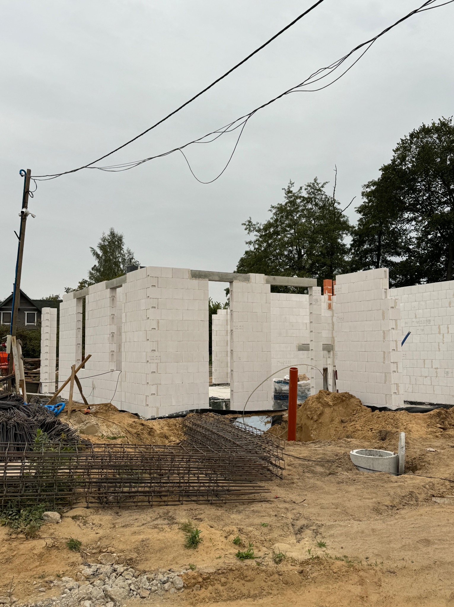 Wznoszone ściany domu z białych bloczków, widoczne zbrojenia i elementy kanalizacyjne na placu budowy, pochmurne niebo w tle.