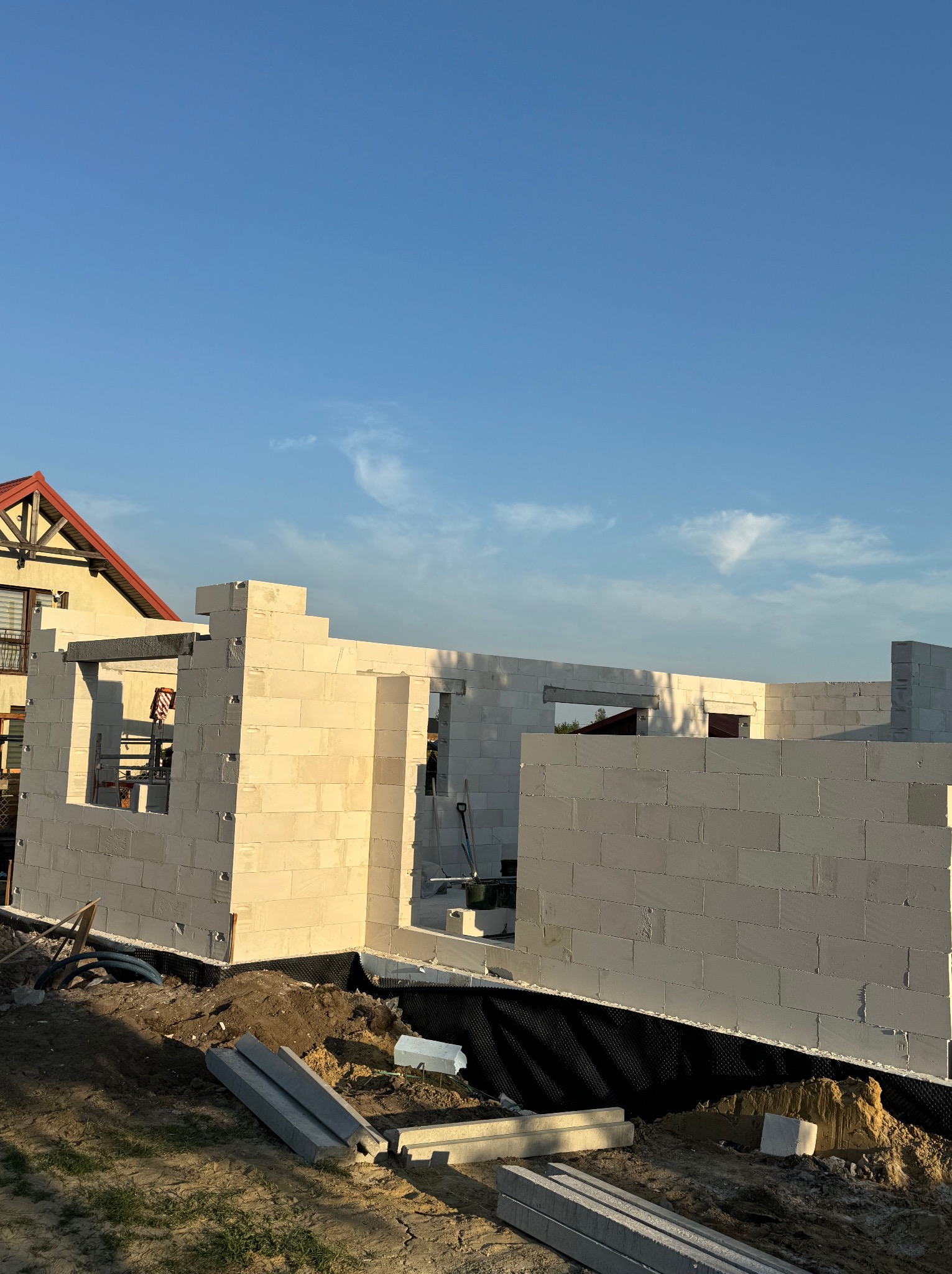 Wznoszenie ścian domu z bloczków betonu komórkowego Ytong w trakcie budowy, widoczne rusztowanie, narzędzia i fragment dachu sąsiedniego budynku w tle.