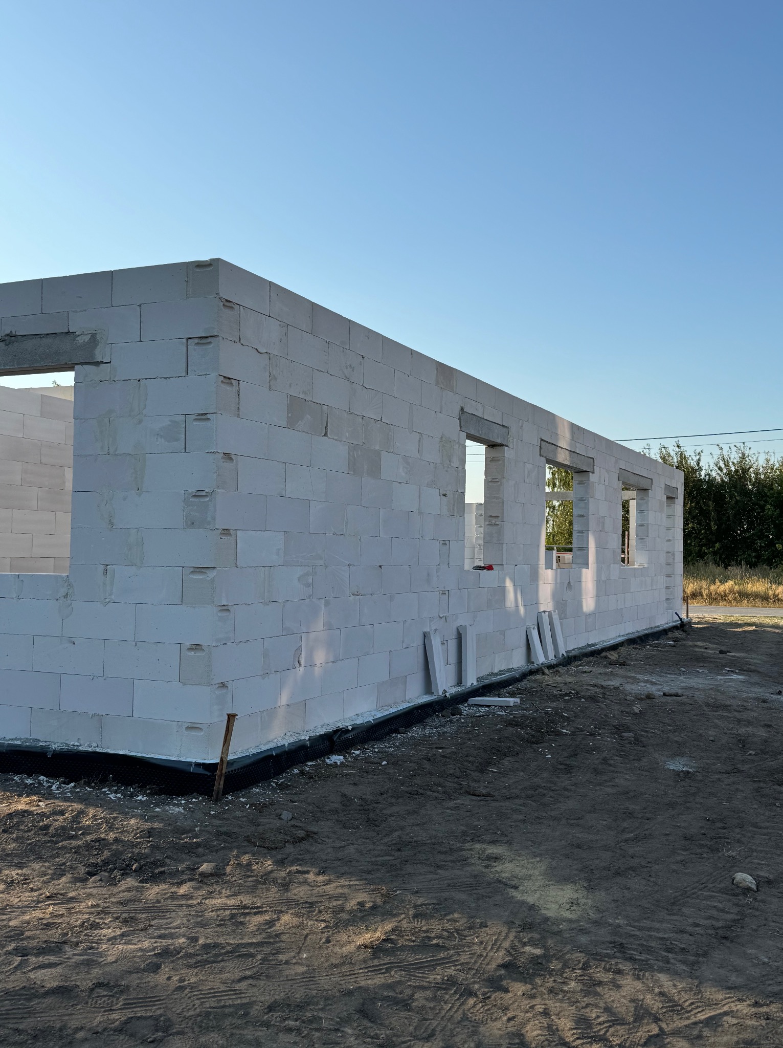 Wznoszenie ścian zewnętrznych budynku z białych bloczków betonowych z widocznymi otworami okiennymi i przygotowanym fundamentem, w słoneczny dzień.