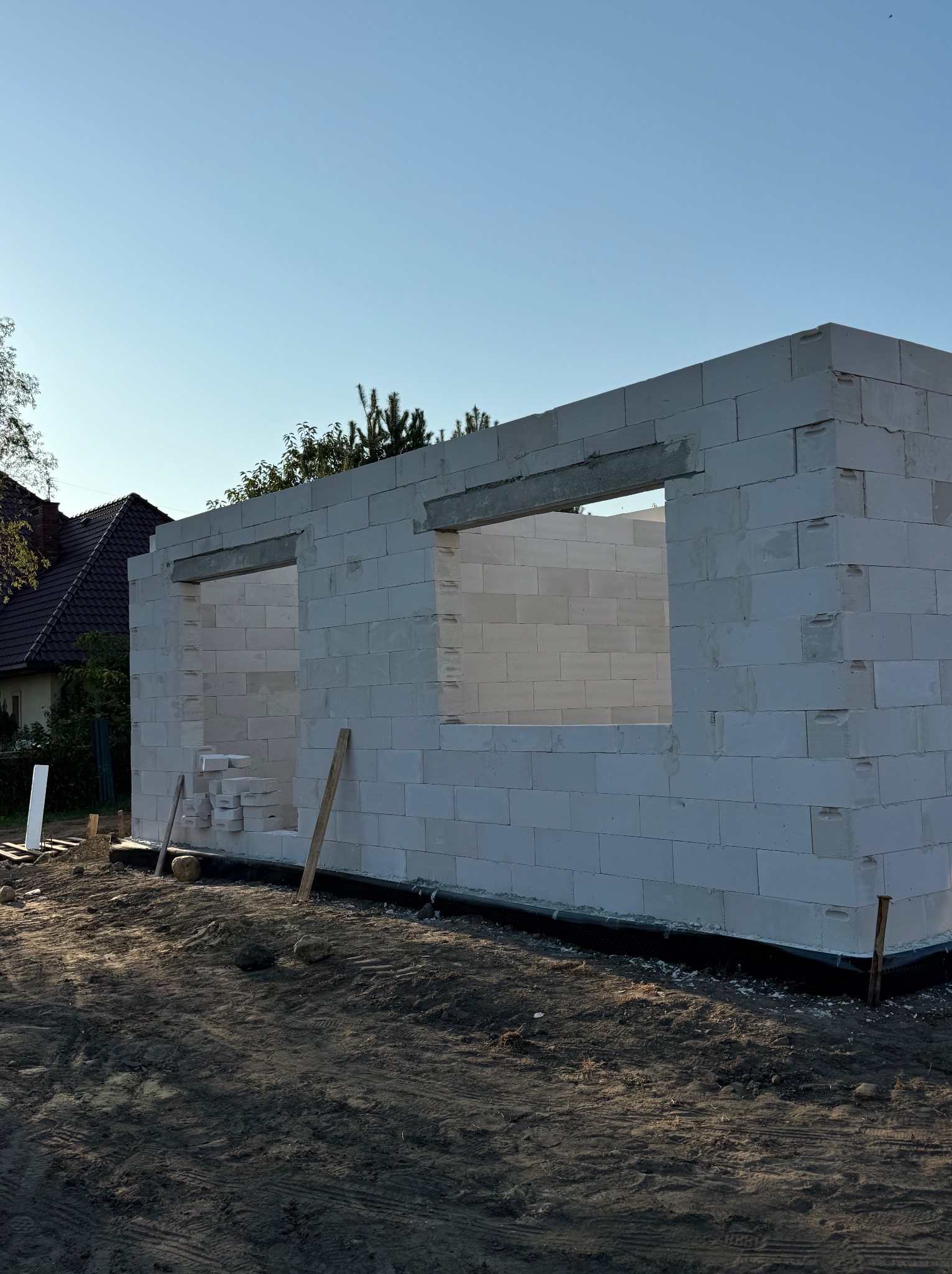 Wznoszona ściana parterowego budynku z betonu komórkowego, widoczne otwory okienne z nadprożami, w tle dom jednorodzinny z ciemnym dachem.