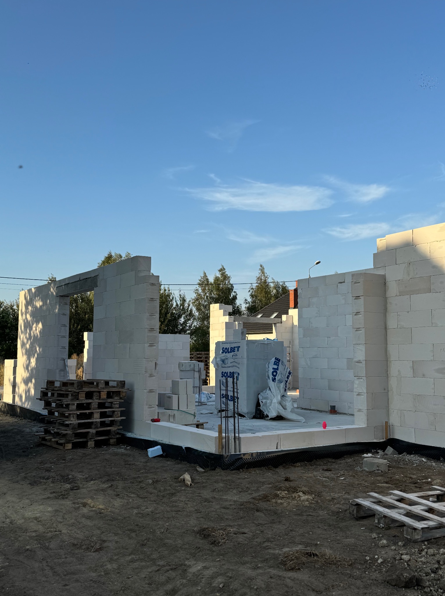 Wznoszenie ścian parterowego domu z białych bloczków betonowych Solbet, widoczne palety z materiałem i zbrojenie wewnątrz fundamentów, błękitne niebo w tle.