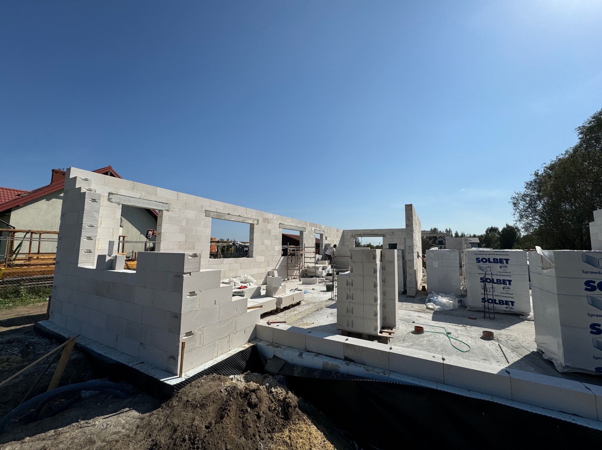 Wznoszone ściany domu z betonu komórkowego Solbet, widoczne palety z bloczkami, fundament zabezpieczony folią kubełkową, słoneczny dzień.