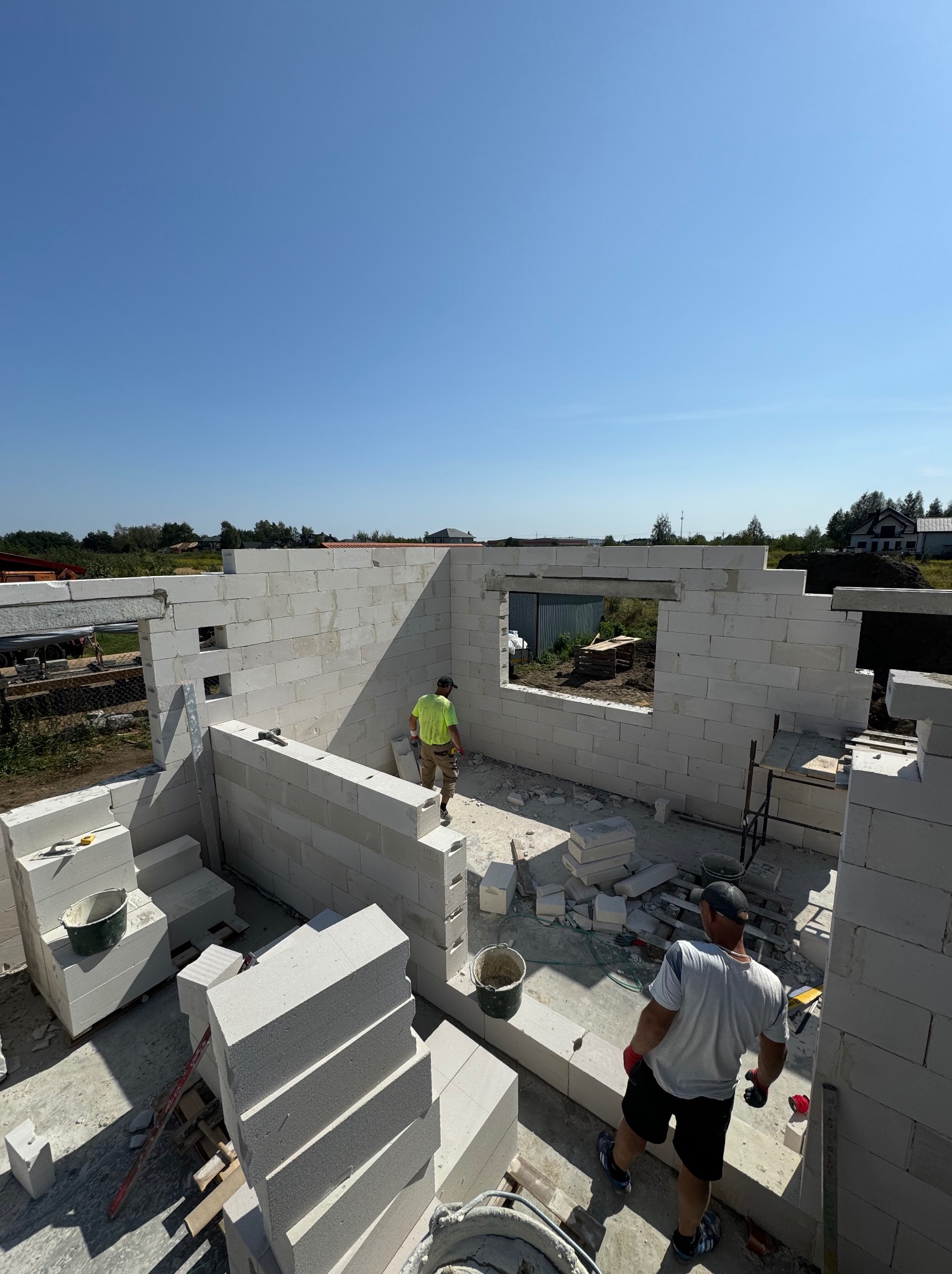 Wznoszenie ścian zewnętrznych domu z betonu komórkowego, widoczni dwaj pracownicy w trakcie pracy, z przodu stos bloczków, w tle widok na okolicę.