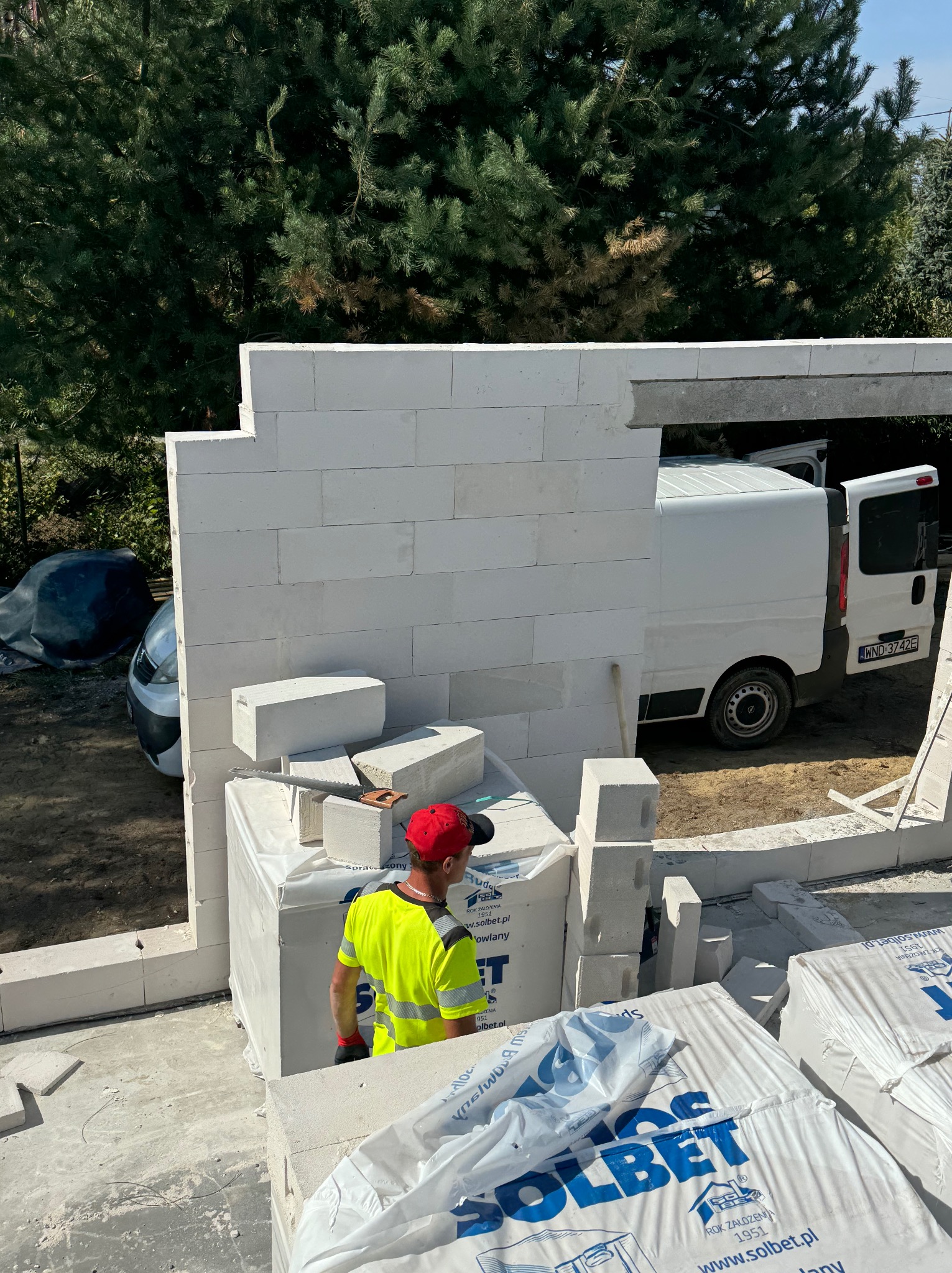 Wznoszenie ściany z białych bloczków Solbet, widoczny pracownik w żółtej kamizelce i czerwonej czapce, paleta z bloczkami i piła ręczna, w tle zaparkowany biały samochód dostawczy.