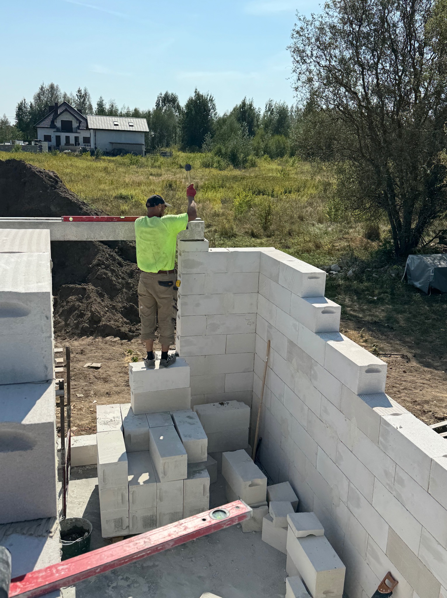Murarz w jaskrawej koszulce i czerwonych rękawicach ustawia bloczki z betonu komórkowego, używając poziomicy, na tle krajobrazu z domem jednorodzinnym.