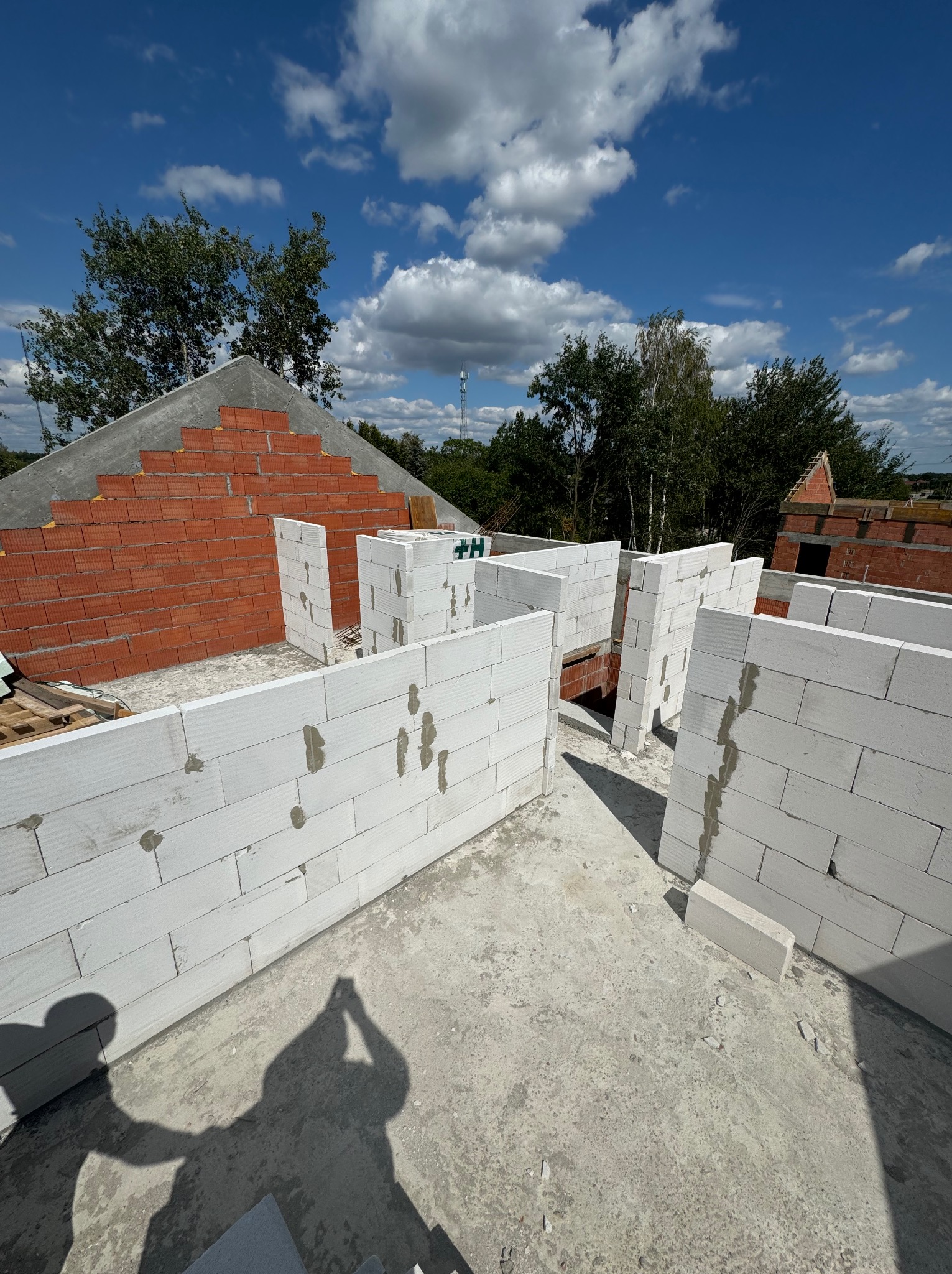 Wznoszenie ścian domu z białych bloczków na fundamencie, widoczny fragment ściany z czerwonej cegły, niebo z chmurami w tle, cień osoby fotografującej w dolnej części kadru.