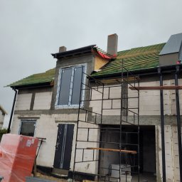 Krzysztof Holak Usługi Remontowo-Budowlane - Budowa domu jednorodzinnego w stanie surowym otwartym, widoczne rusztowania, częściowo pokryty dach z zieloną dachówką, komin, zamontowane okna i drzwi wejściowe, na pierwszym planie materiały...