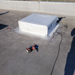 Krzysztof Holak Usługi Remontowo-Budowlane - Szary dach płaski z obróbką komina w białej membranie, widoczny wiertarko-wkrętak akumulatorowy, opalarka dekarska i wkrętak płaski, w tle zabudowania przemysłowe