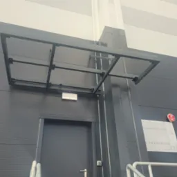 Szara metalowa konstrukcja daszku nad wejściem do budynku o panelowej elewacji, widoczne elementy instalacji przeciwpożarowej.