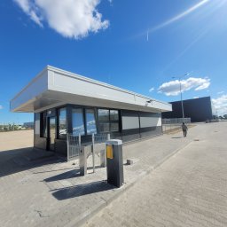 Krzysztof Holak Usługi Remontowo-Budowlane - Nowoczesny, biało-czarny budynek portierni z płaskim dachem, widoczny z zewnątrz na tle błękitnego nieba i placu manewrowego.