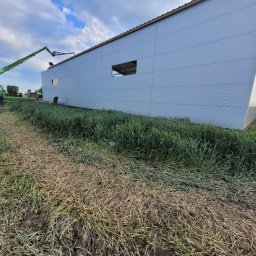 Krzysztof Holak Usługi Remontowo-Budowlane - Montaż paneli ściennych na budynku przemysłowym, widoczny podnośnik koszowy i pracownicy na dachu, niezagospodarowany teren wokół budynku.