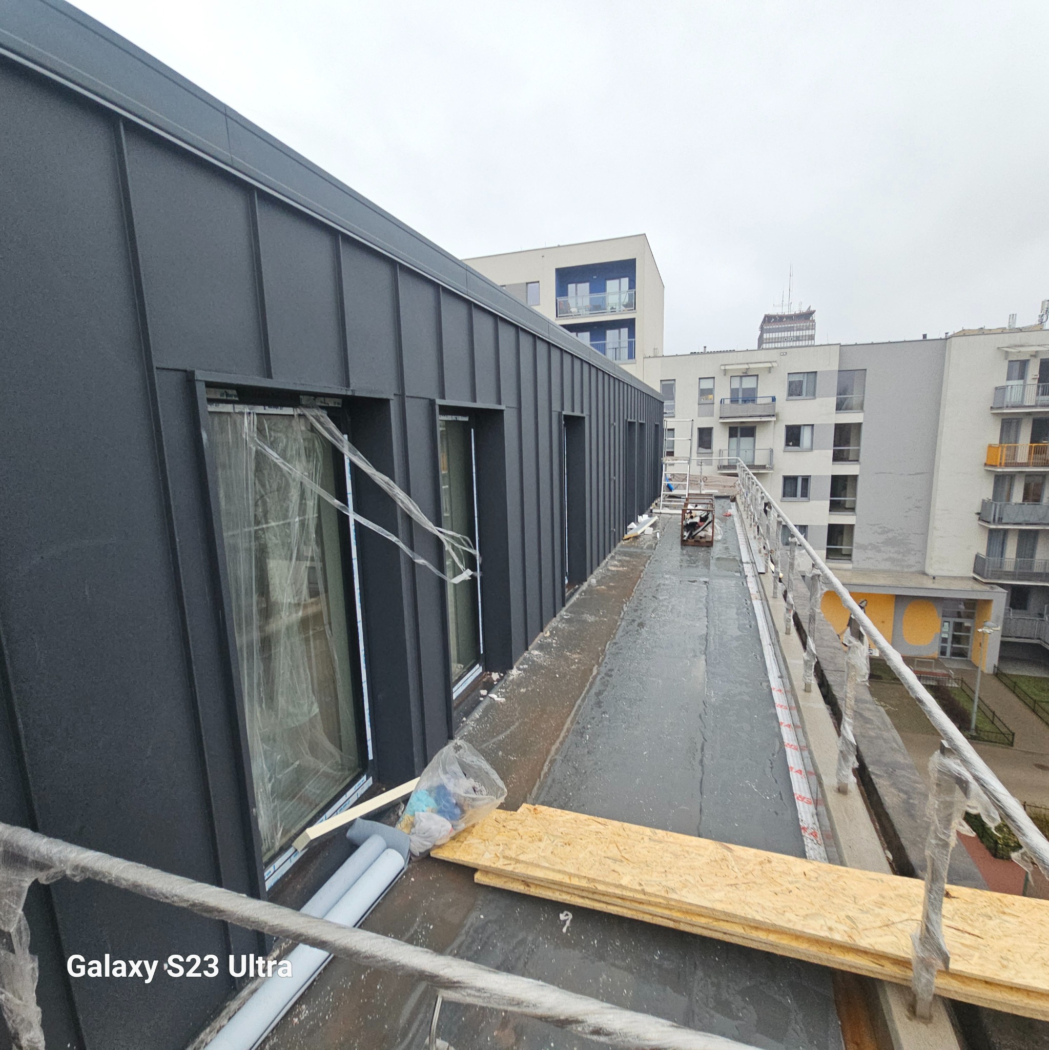 Nowoczesna elewacja budynku z ciemnymi panelami, widoczne okna z folią ochronną, materiały budowlane na dachu oraz budynki w tle w pochmurny dzień.