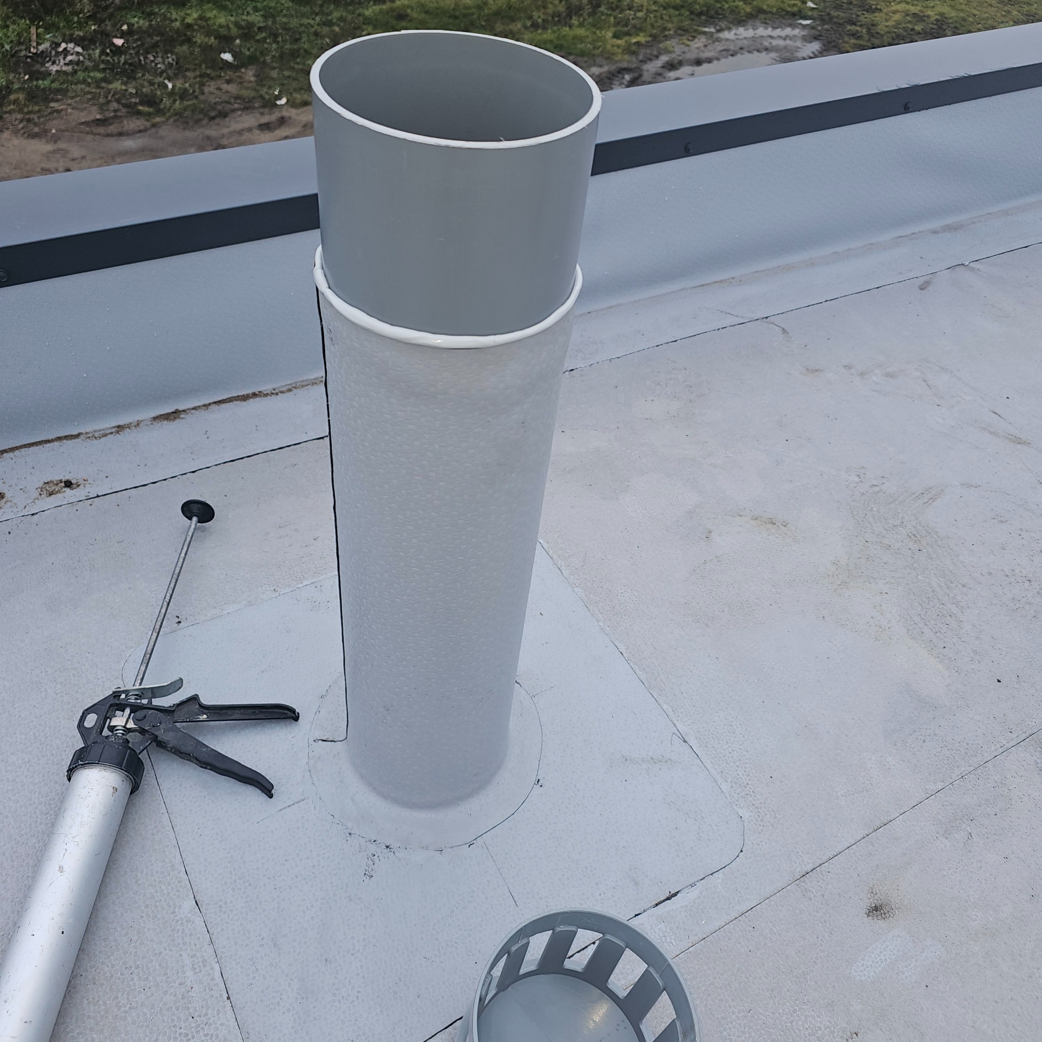 Szary kominek wentylacyjny na płaskim dachu, uszczelniony silikonem, z pistoletem do silikonu obok i fragmentem obudowy kominka.