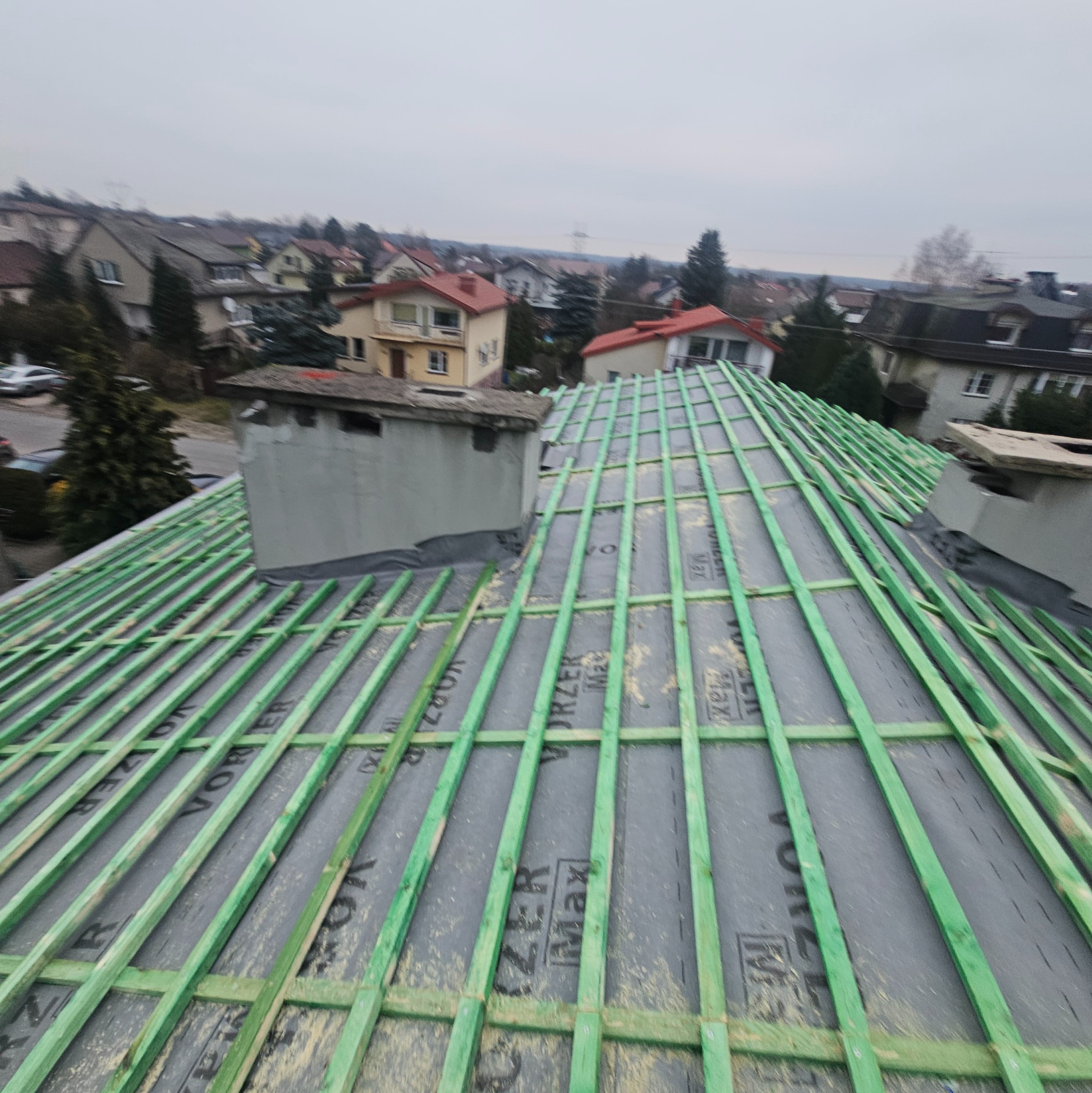 Konstrukcja dachu z zielonych łat na czarnej membranie, widok z góry na tle zabudowań miejskich i pochmurnego nieba.