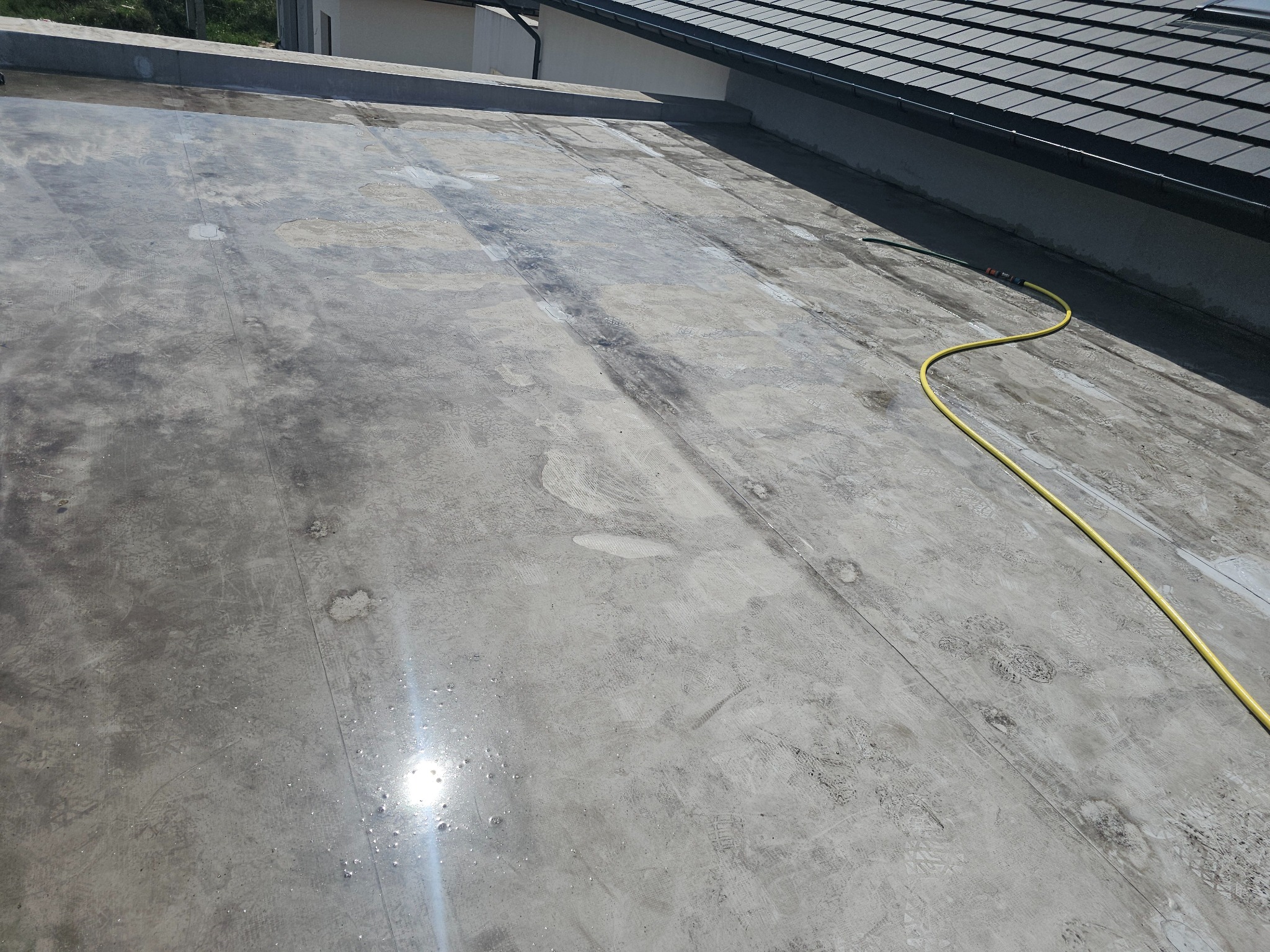 Płaski dach pokryty papą z widocznymi śladami użytkowania i żółtym wężem ogrodowym wzdłuż krawędzi, w tle dach budynku z szarej dachówki.