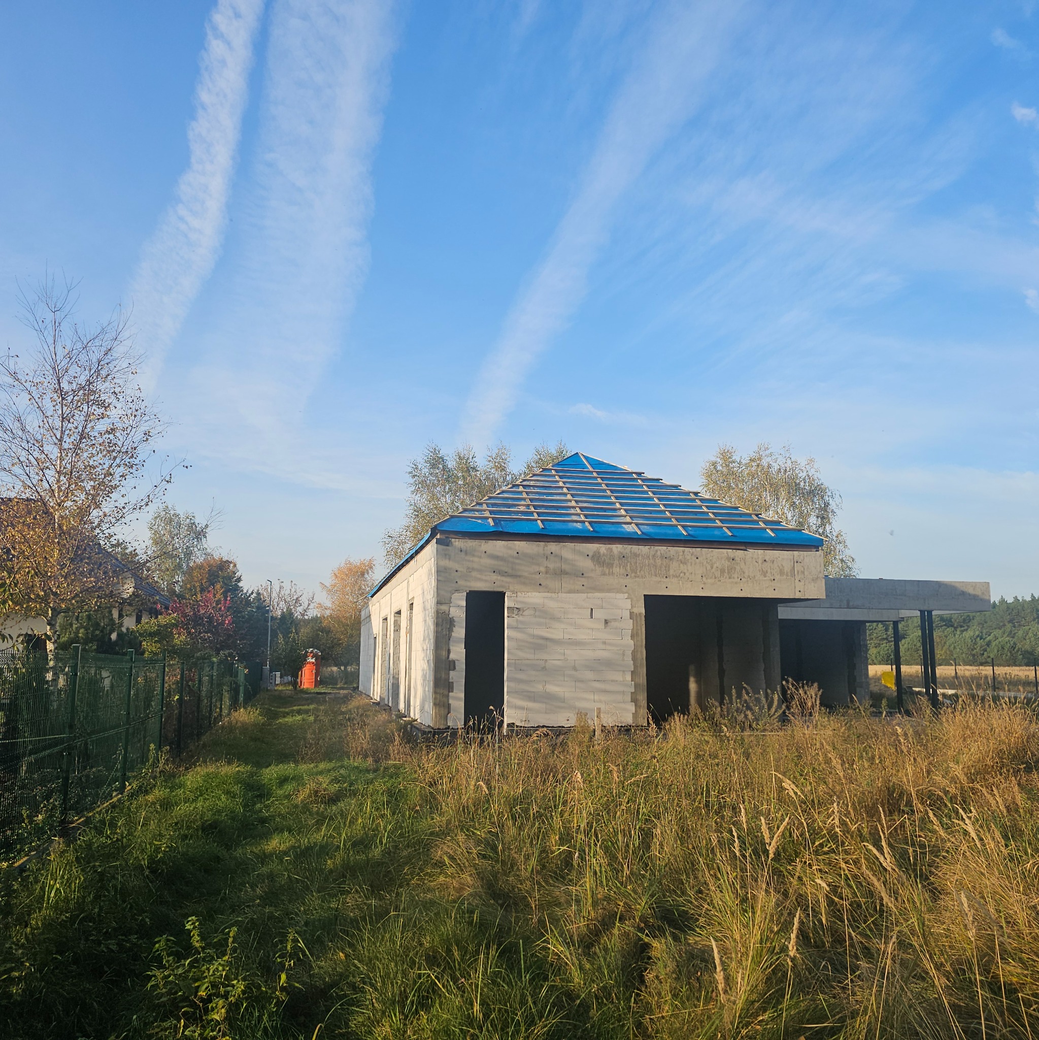 Surowy budynek w trakcie budowy z niebieską folią na drewnianej więźbie dachowej, otoczony wysoką trawą i drzewami, na tle błękitnego nieba z smugami kondensacyjnymi.