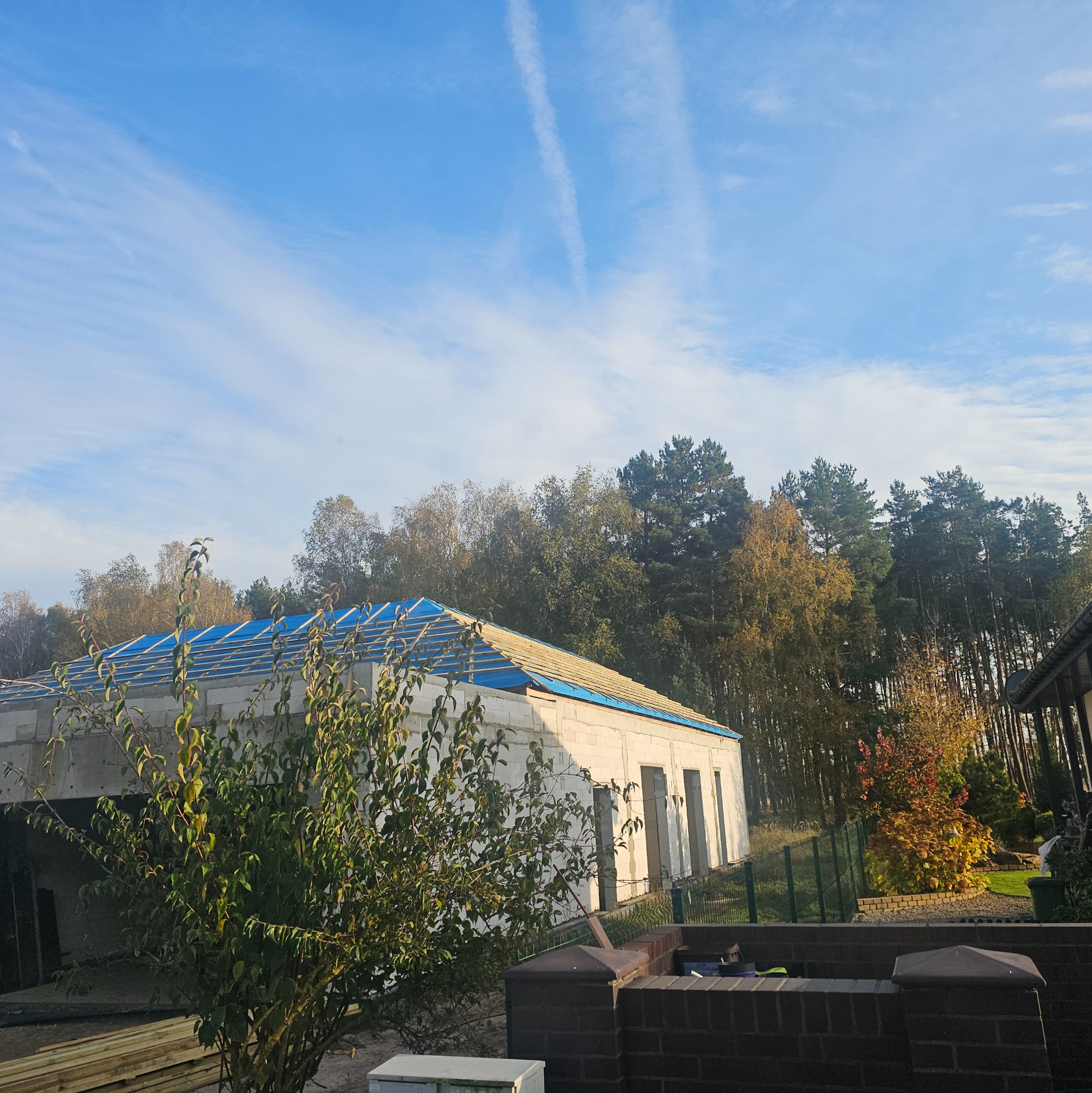 Budynek w trakcie budowy z częściowo zamontowaną konstrukcją dachu, zabezpieczoną niebieską folią, na tle lasu i błękitnego nieba z smugami kondensacyjnymi.