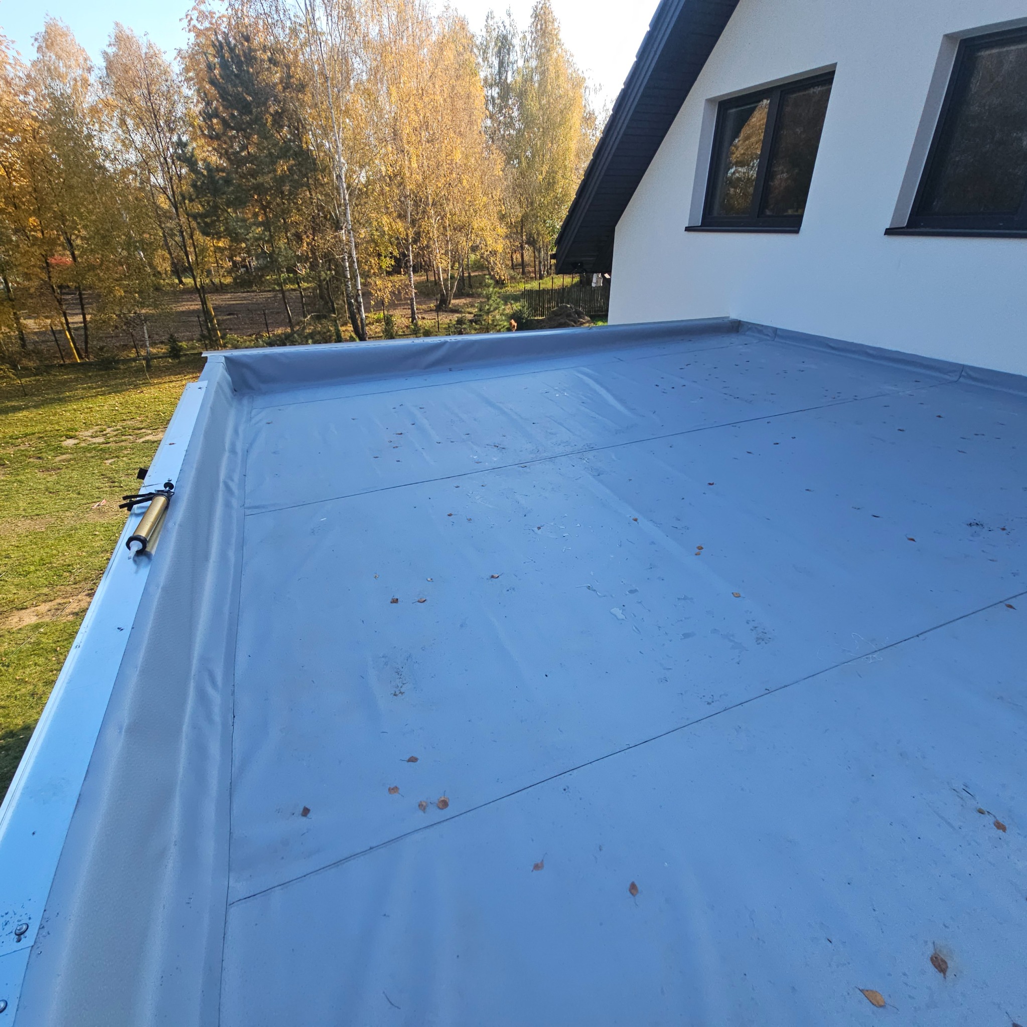 Płaski dach pokryty szarą membraną, widoczny pistolet do uszczelniania na krawędzi, w tle biała ściana budynku z czarnymi oknami i jesienne drzewa.