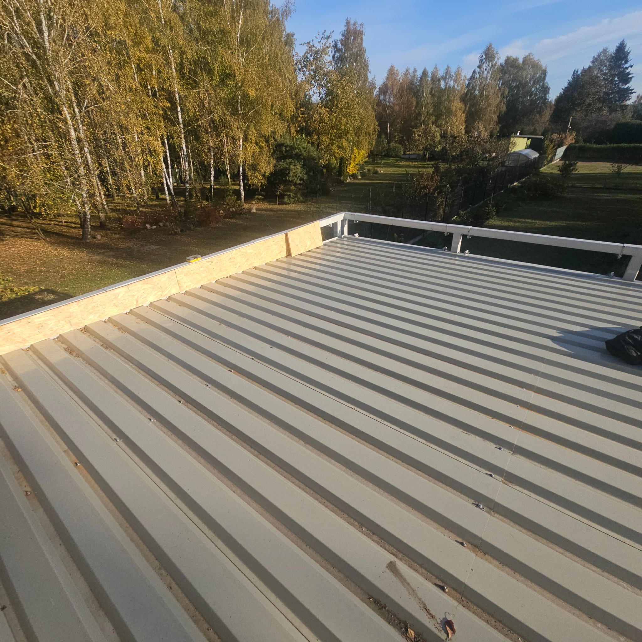 Nowy, szary dach z blachy trapezowej w trakcie montażu, widoczne deski okapowe i balustrada, w tle drzewa w jesiennych barwach.