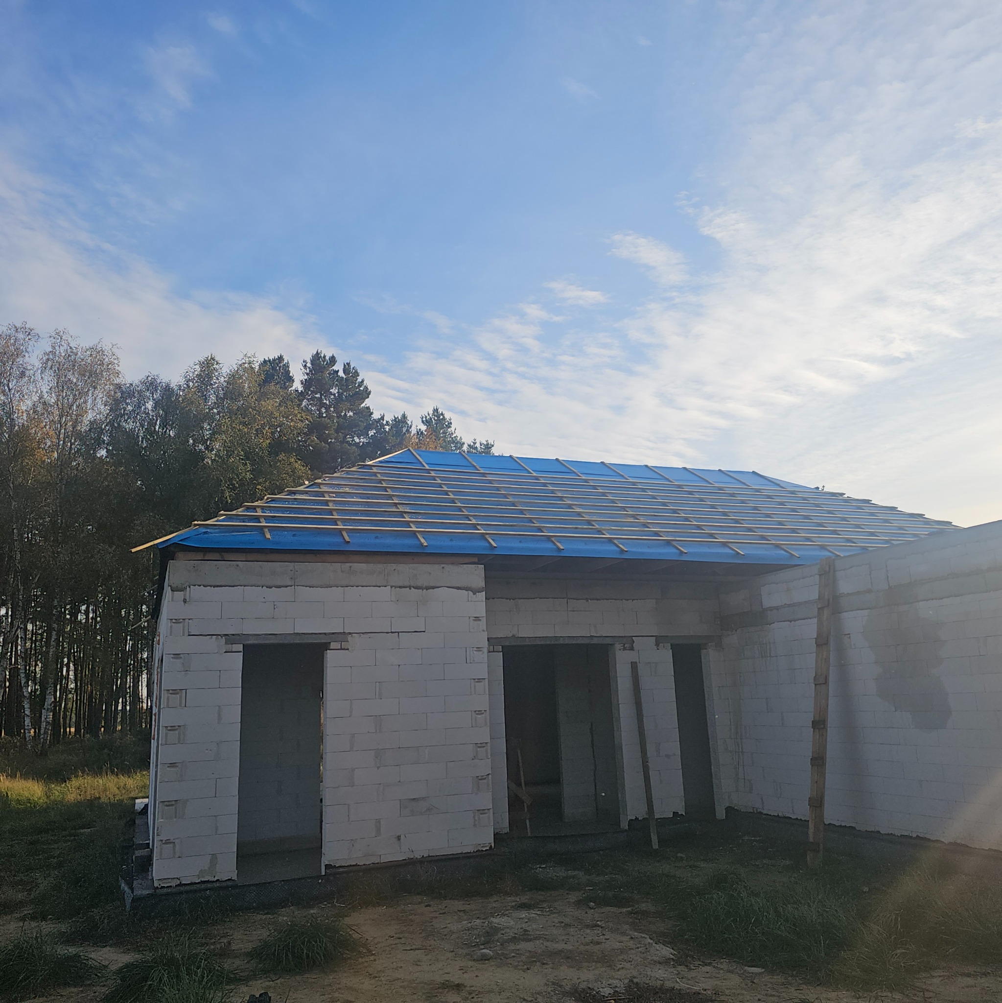 Budowa domu jednorodzinnego w stanie surowym otwartym z niebieską folią na drewnianej konstrukcji dachu, widok z zewnątrz w słoneczny dzień.