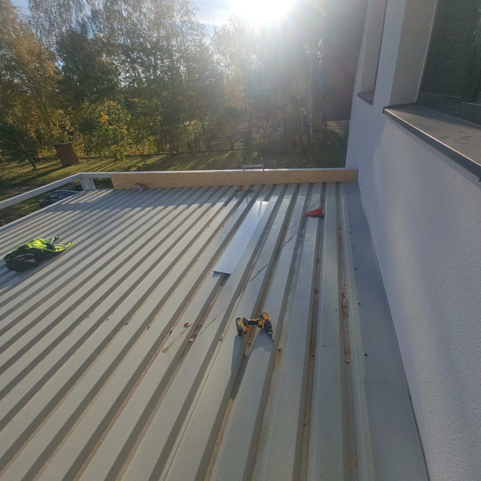 Szara blacha trapezowa na dachu domu jednorodzinnego w trakcie montażu, widoczne narzędzia, fragment obróbki blacharskiej i elementy mocujące, w tle drzewa i fragment elewacji.