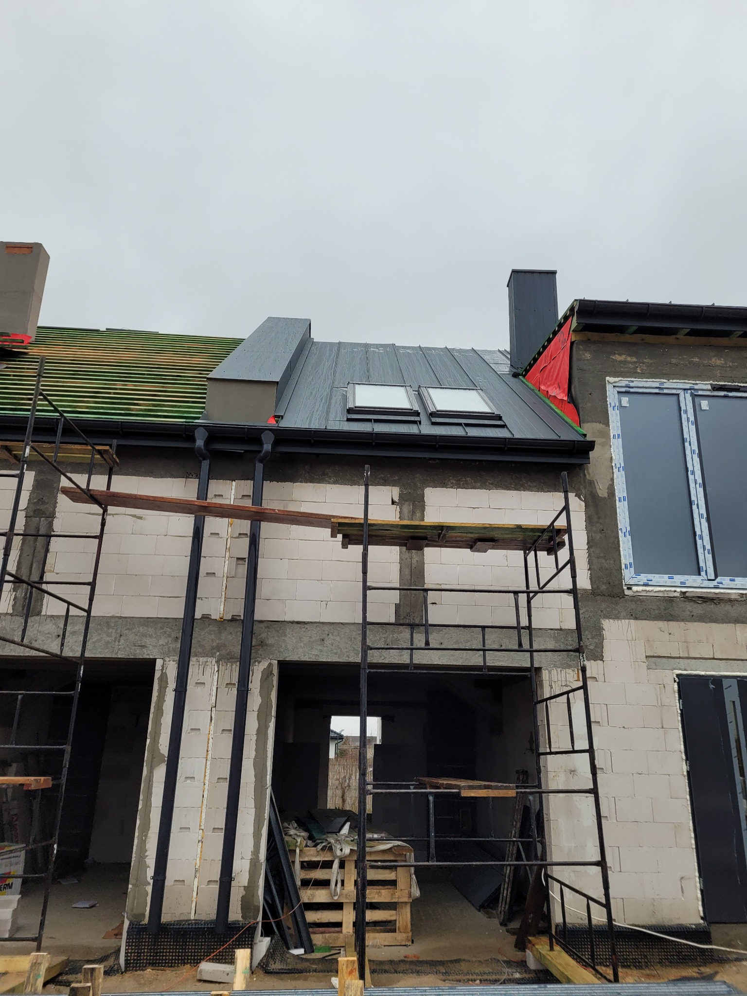 Budynek w trakcie budowy z widocznymi elementami dachu: fragment zielonej konstrukcji, szara blacha z oknami dachowymi, czarne rynny oraz rusztowania.