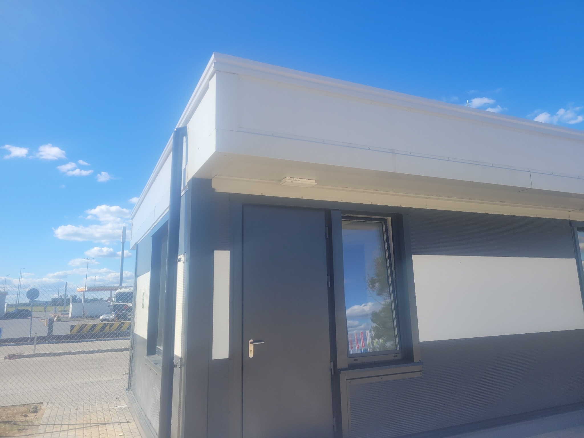 Nowoczesny budynek z płaskim dachem i metalową elewacją, widoczne okno z odbiciem nieba.