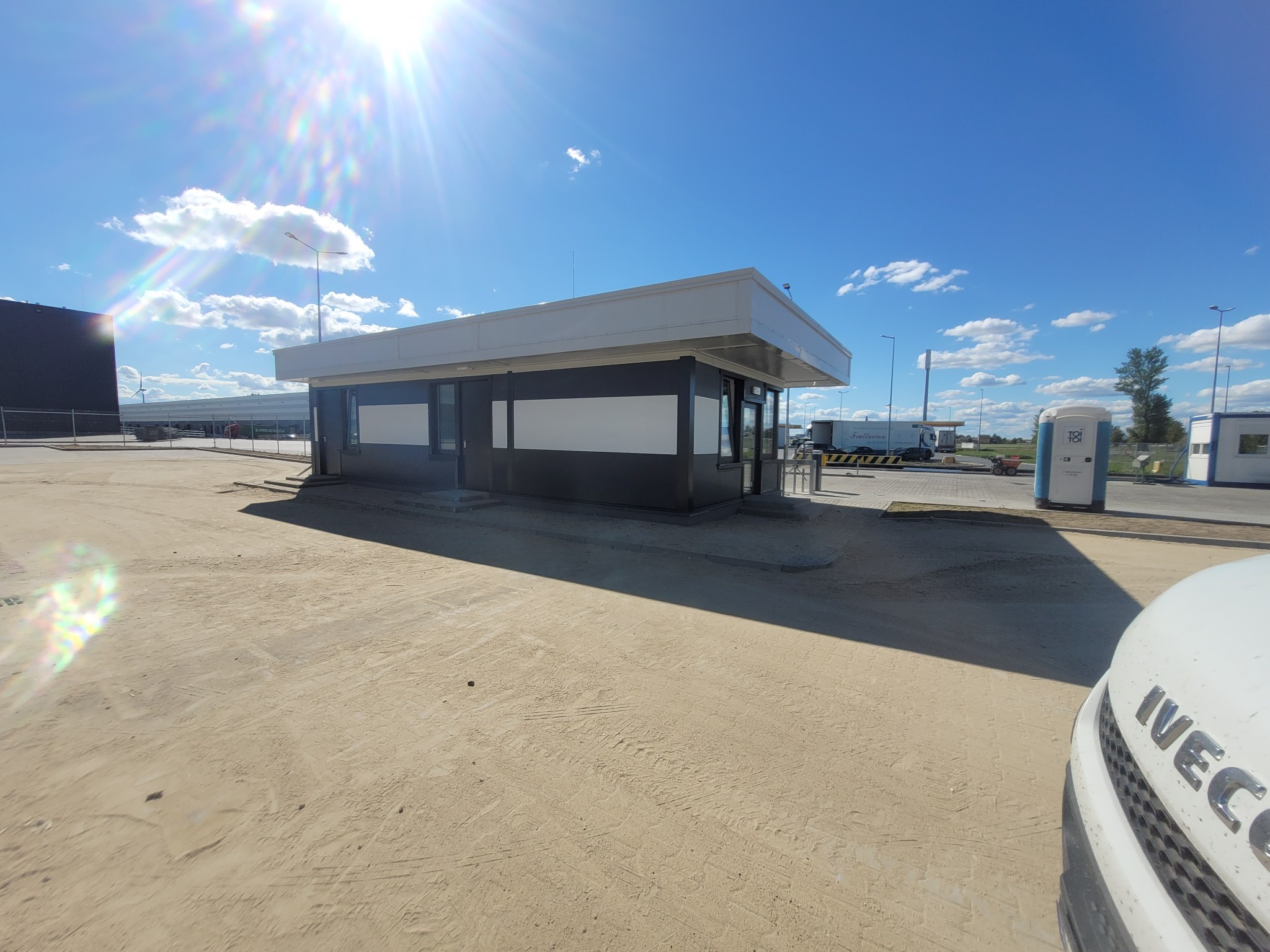 Nowoczesny, parterowy budynek biurowy z płaskim dachem, pomalowany w kolorach szaro-białych, usytuowany na placu budowy z widocznym pojazdem Iveco w prawym dolnym rogu i przenośną toaletą w tle,...